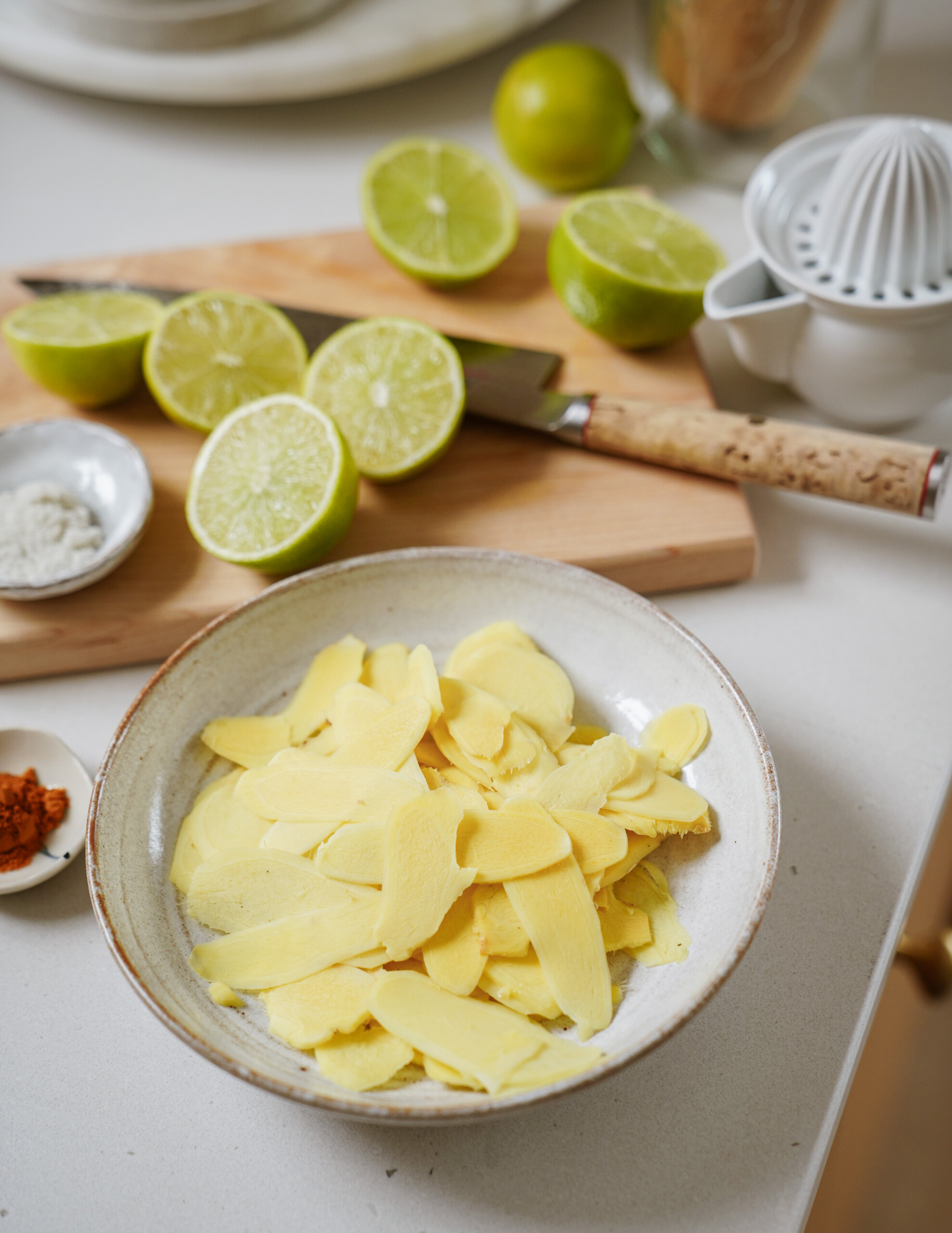 A bowl of thinly sliced ginger sits on a counter beside a cutting board with halved limes, a knife, a citrus juicer, and small spice dishes.