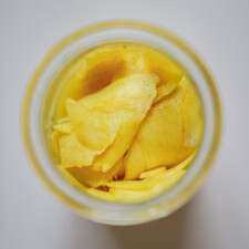 A top-down view of thin yellow mango slices in a glass jar, with a white background.