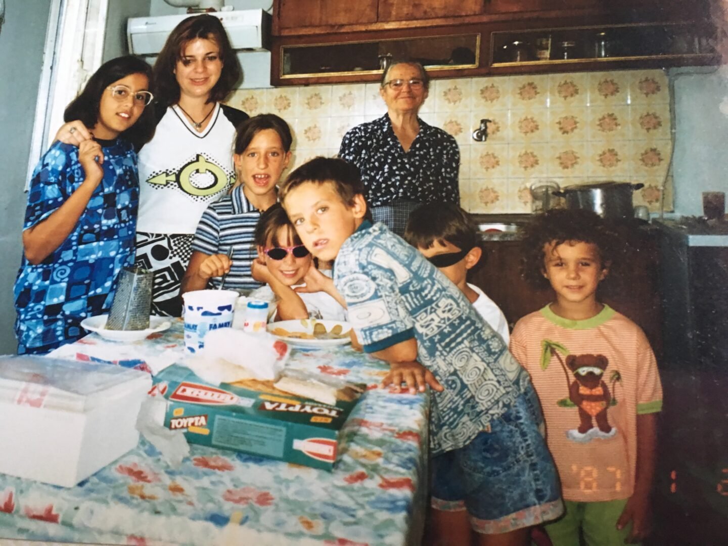 A group of children and two adults smile in a kitchen, gathered around a table covered with food and kitchen items. The kitchen features patterned tiles and wooden cabinets.