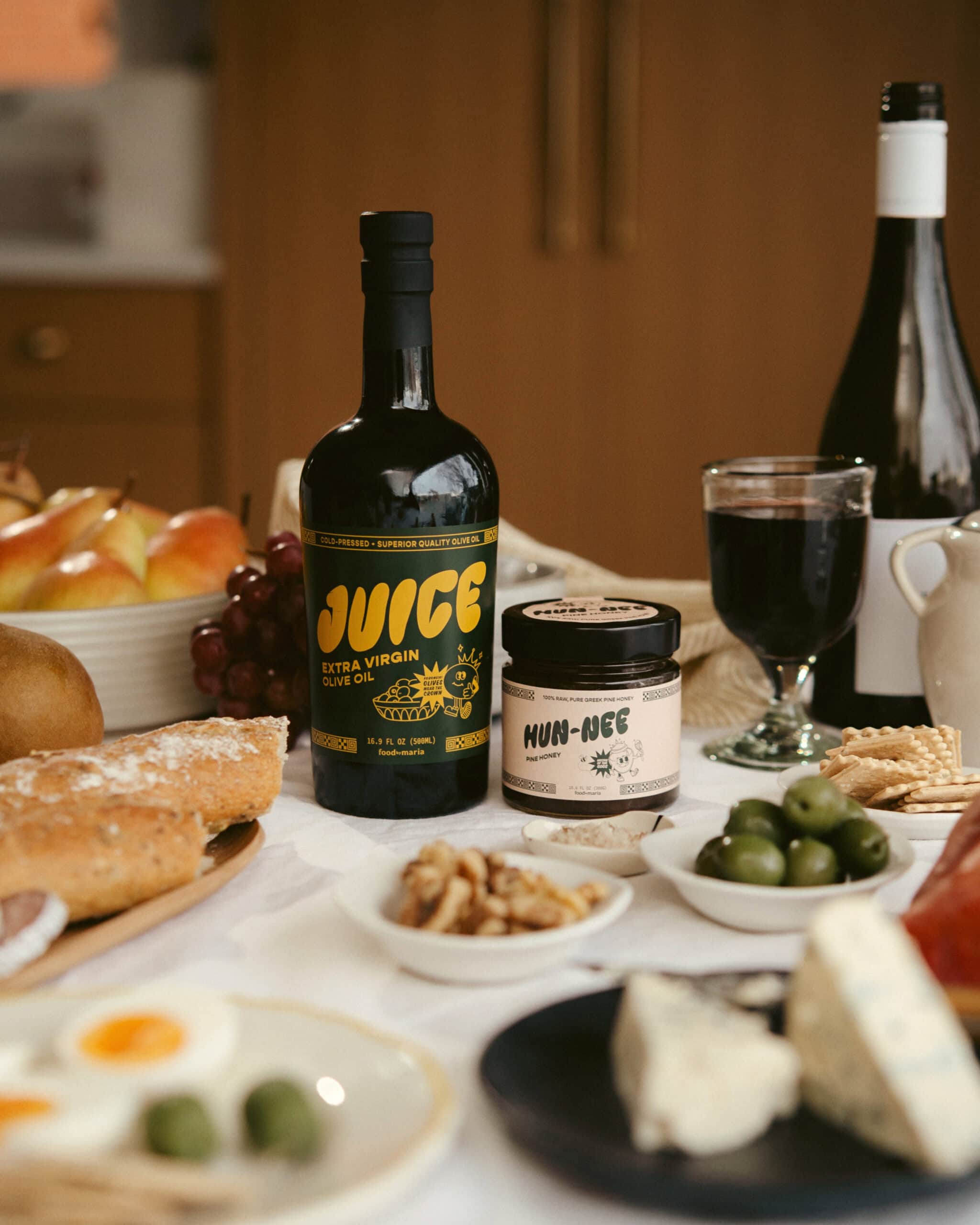 A table set with a bottle of olive oil labeled Juice, a jar of Hun-Nec, wine, a glass of red wine, bread, cheese, hard-boiled eggs, grapes, walnuts, olives, apples, and crackers.