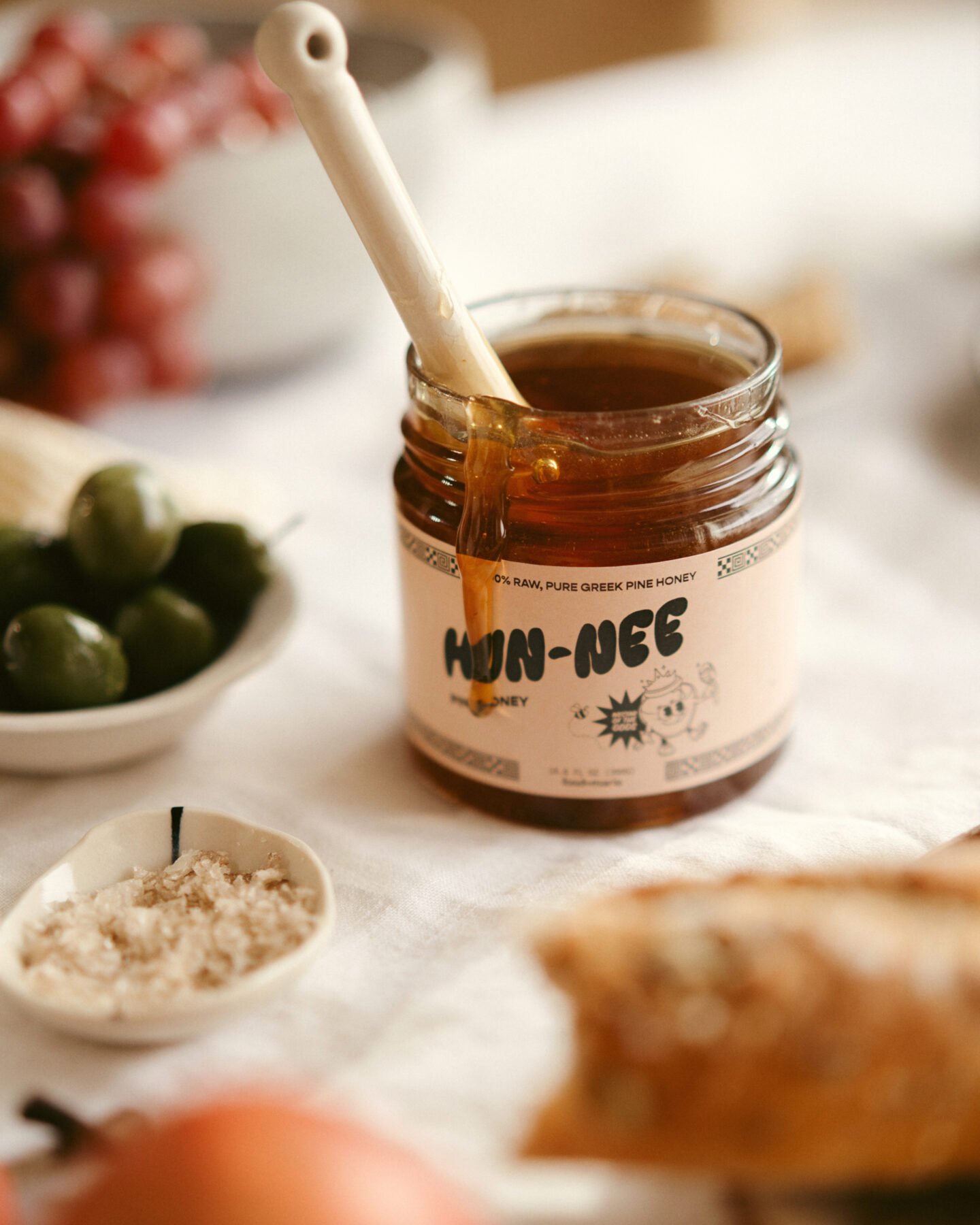 A jar of honey labeled HUN-NEE with a ceramic spoon inside sits on a table surrounded by olives, grapes, bread, and a small dish of salt.