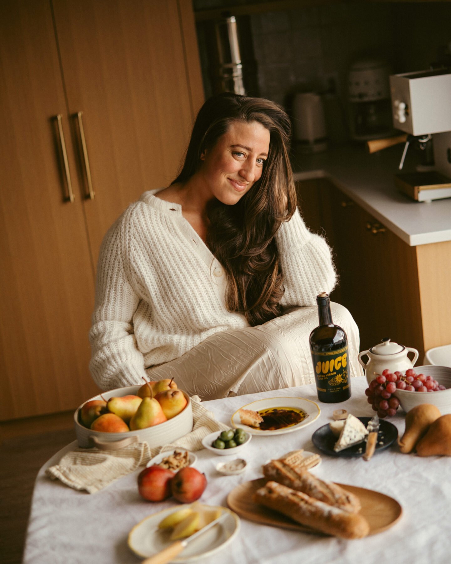 A woman in a white sweater sits at a table with pears, grapes, pomegranate, bread, cheese, olives, olive oil, and a bottle of wine, in a cozy kitchen setting with wooden cabinets.