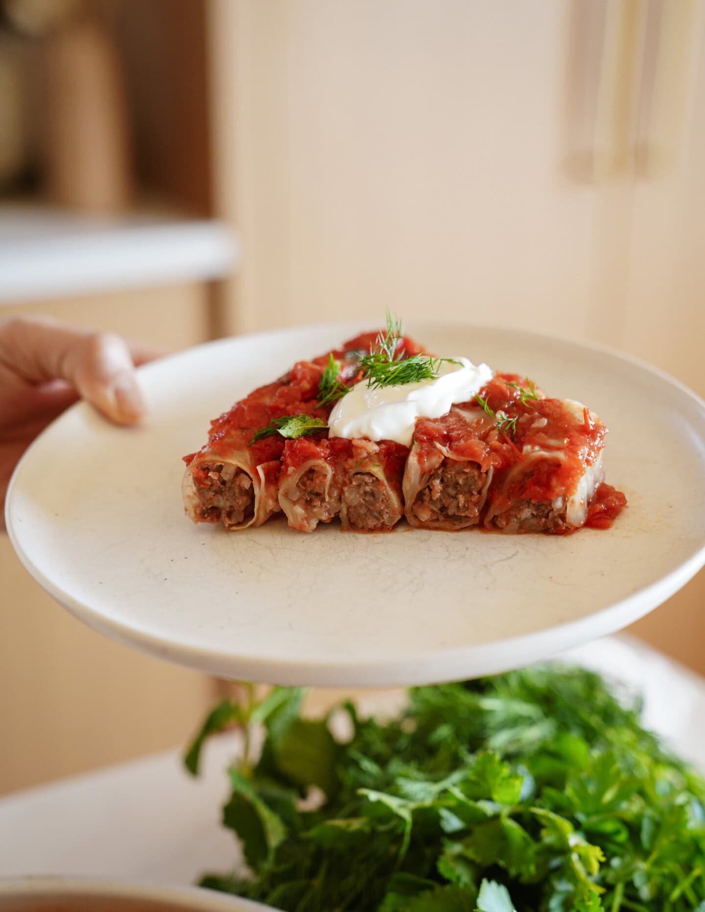 A plate with five pieces of stuffed rolled crepes topped with tomato sauce and a dollop of sour cream, garnished with fresh herbs. In the background, there are fresh green herbs on a table.