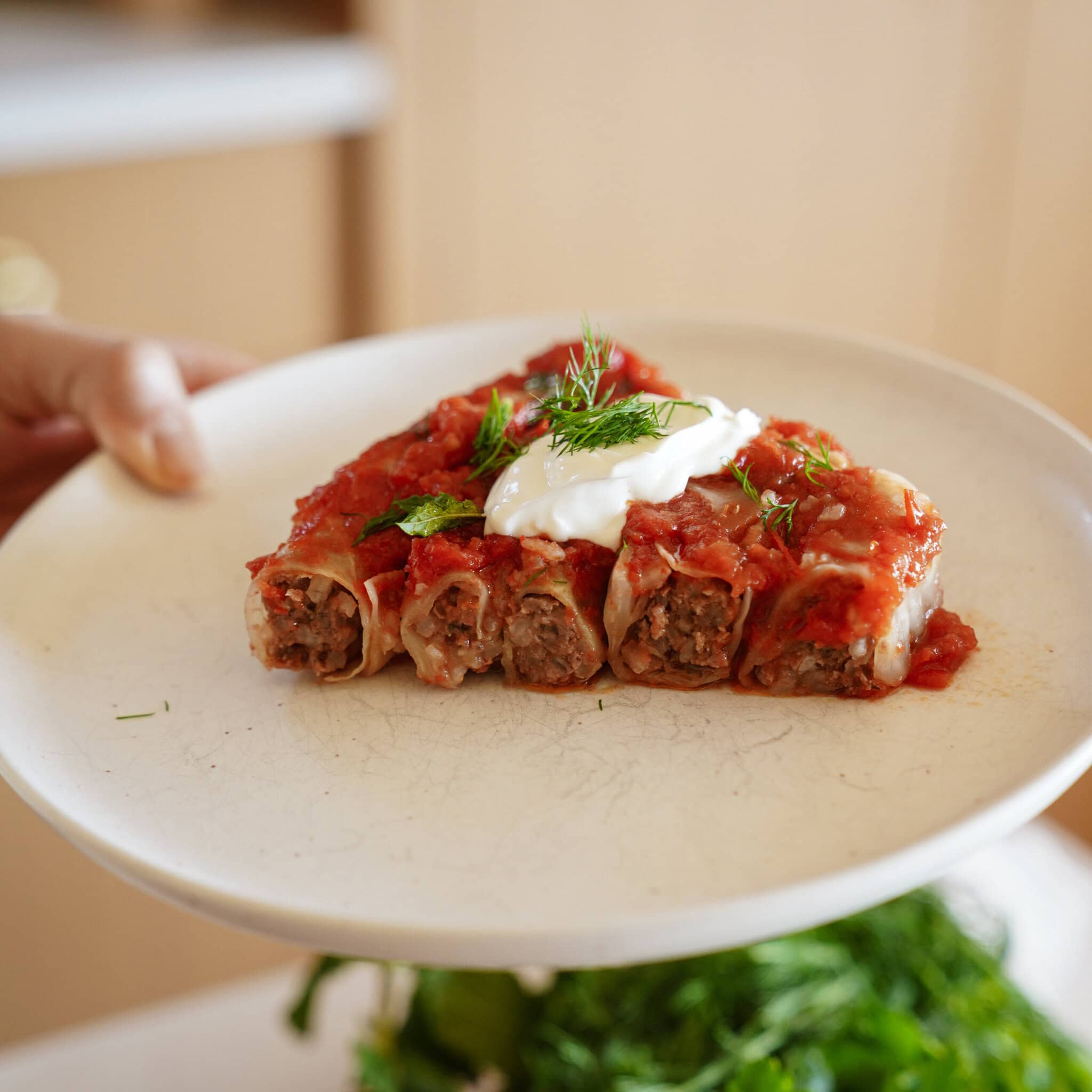 A white plate with five pieces of meat-stuffed cabbage rolls topped with tomato sauce, a dollop of sour cream, and sprigs of fresh dill, held by a hand in a kitchen setting.