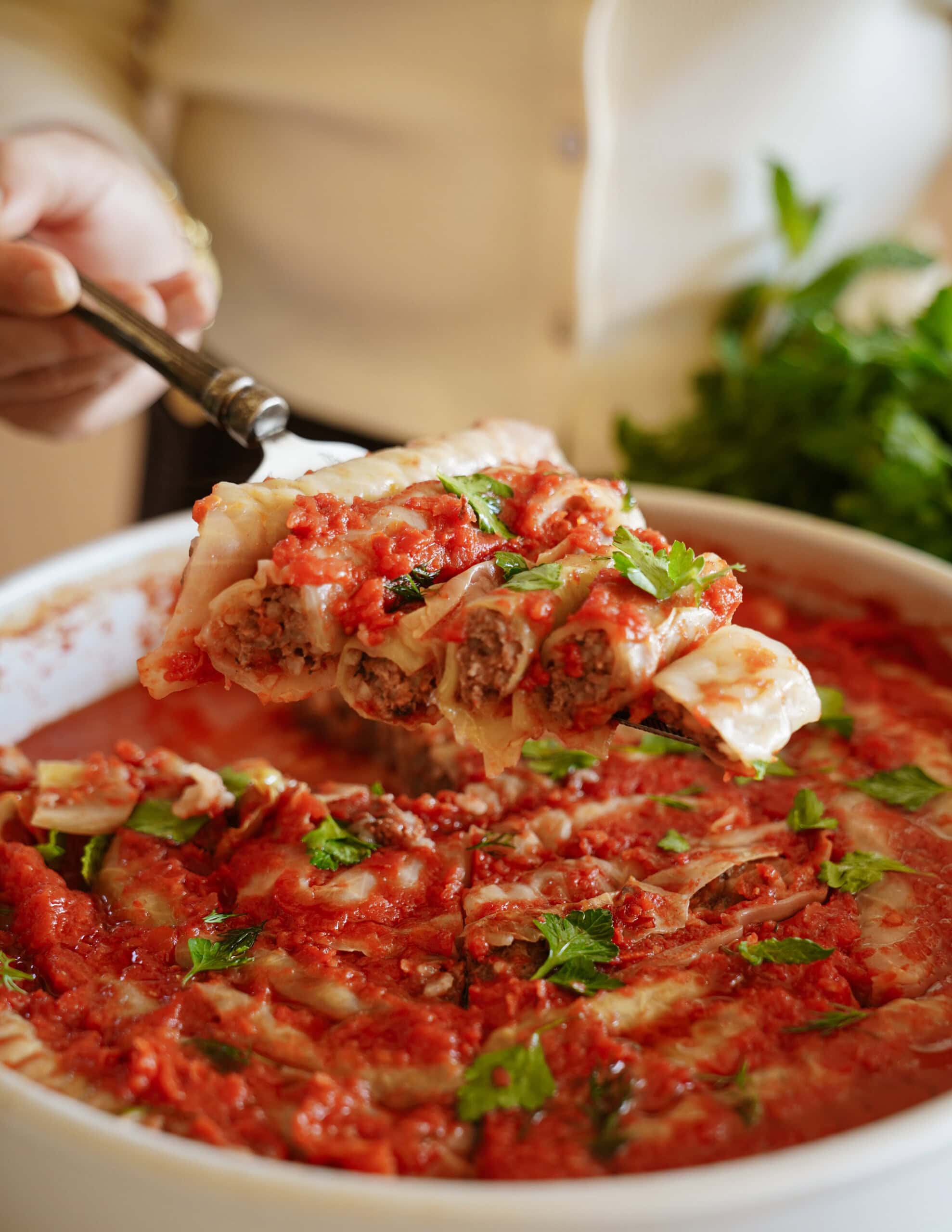 A hand serving a portion of stuffed cabbage rolls topped with tomato sauce and parsley from a white baking dish, with fresh herbs in the background.
