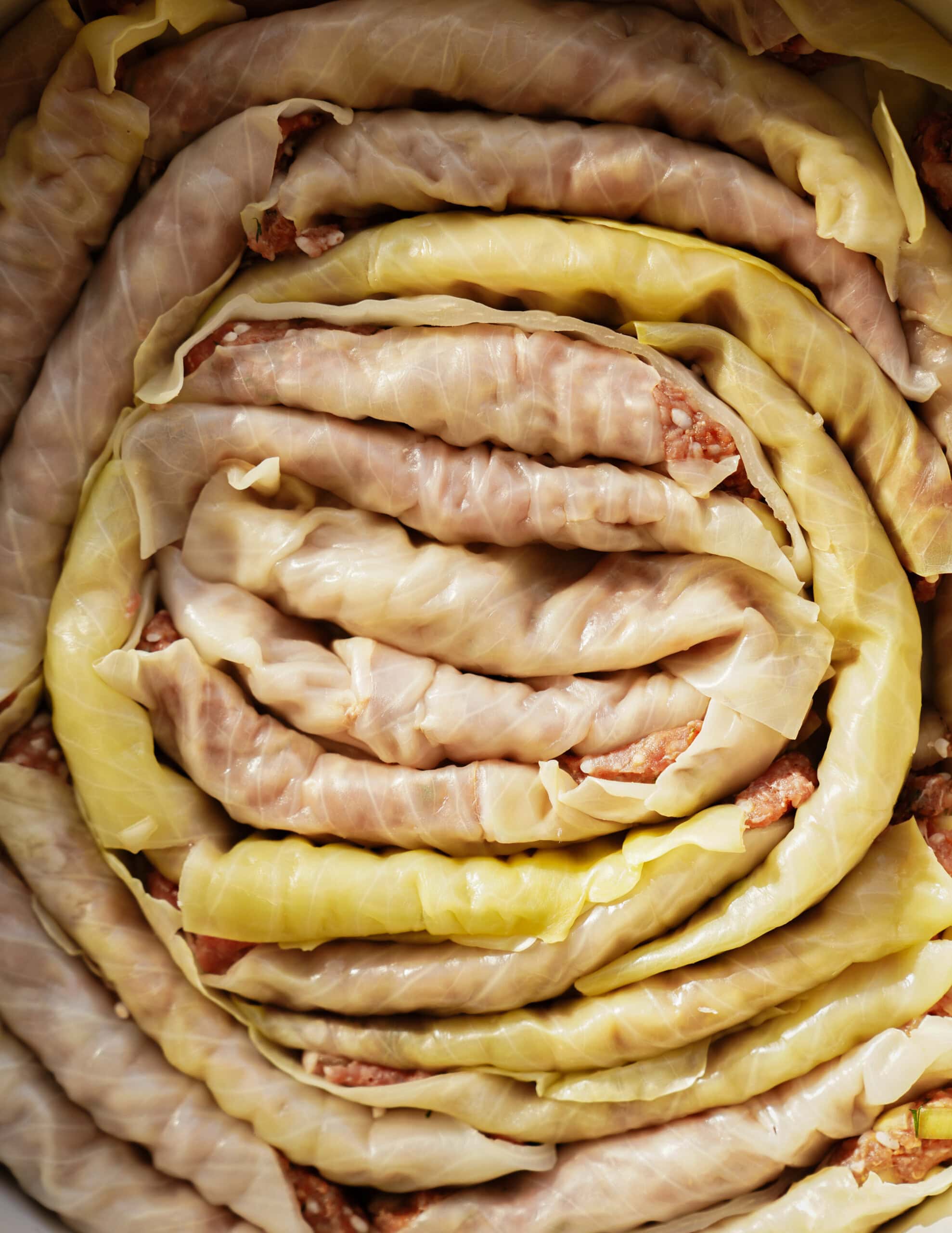A close-up of raw cabbage rolls filled with ground meat, tightly packed and arranged in a spiral pattern inside a pot. The cabbage leaves appear slightly translucent and light green.