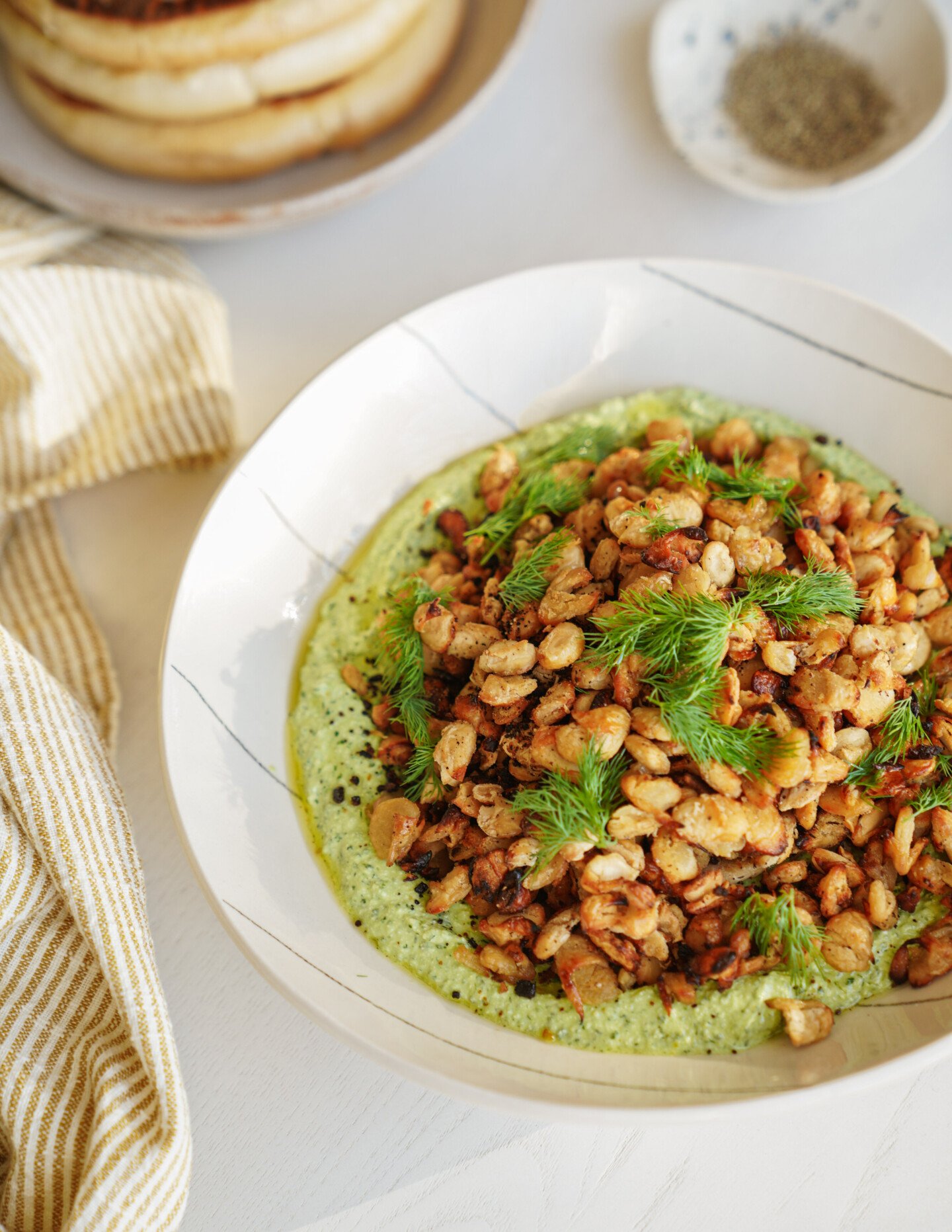 A white bowl filled with sautéed white beans and fresh dill on top of a creamy green sauce, placed on a white table with a striped cloth and other dishes partially visible in the background.