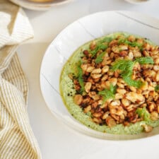 A bowl of crispy roasted beans topped with fresh dill, served over a creamy green sauce, sits on a white table next to a striped beige and white cloth.