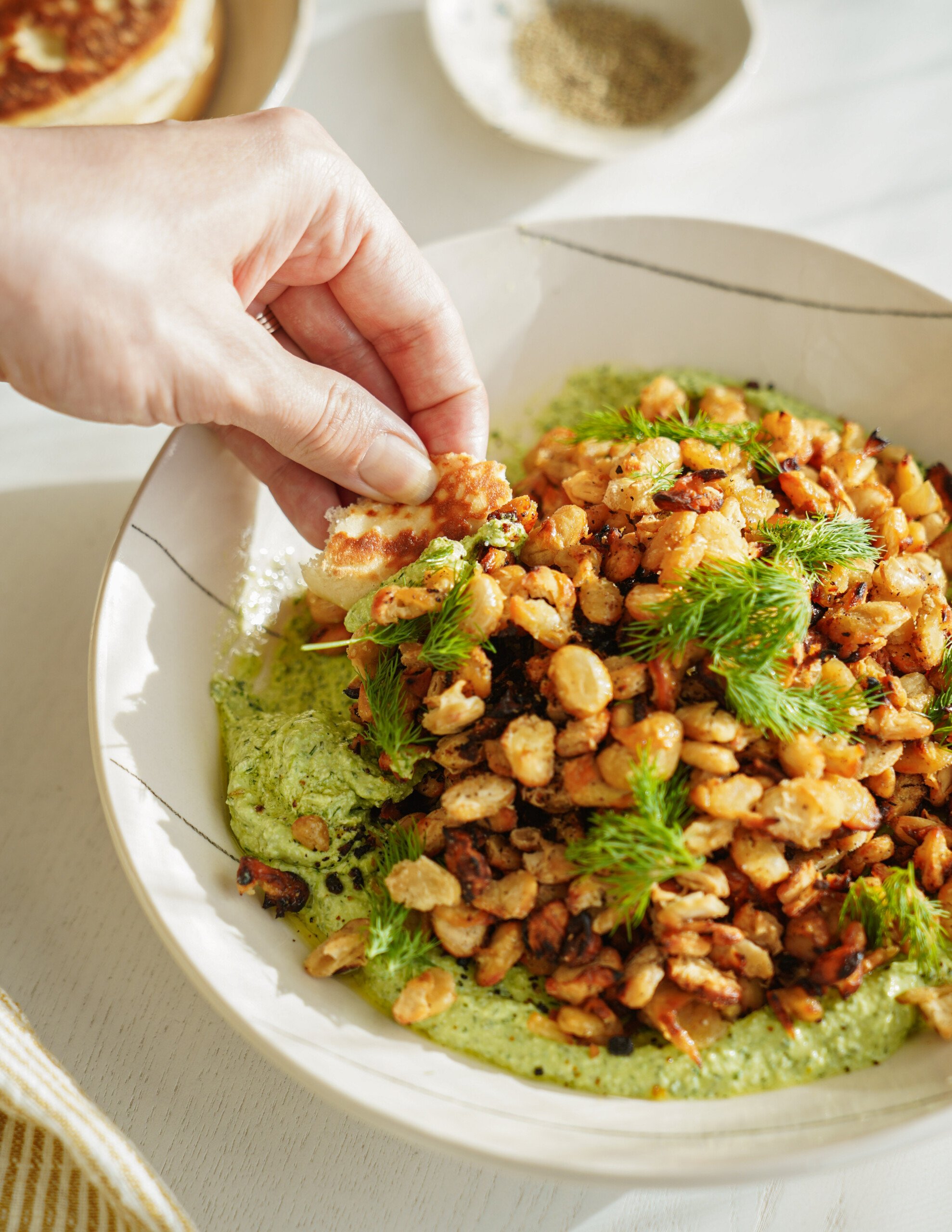 A hand dips a piece of food into a bowl filled with crispy chickpeas, fresh herbs, and a green creamy sauce, set on a light table.