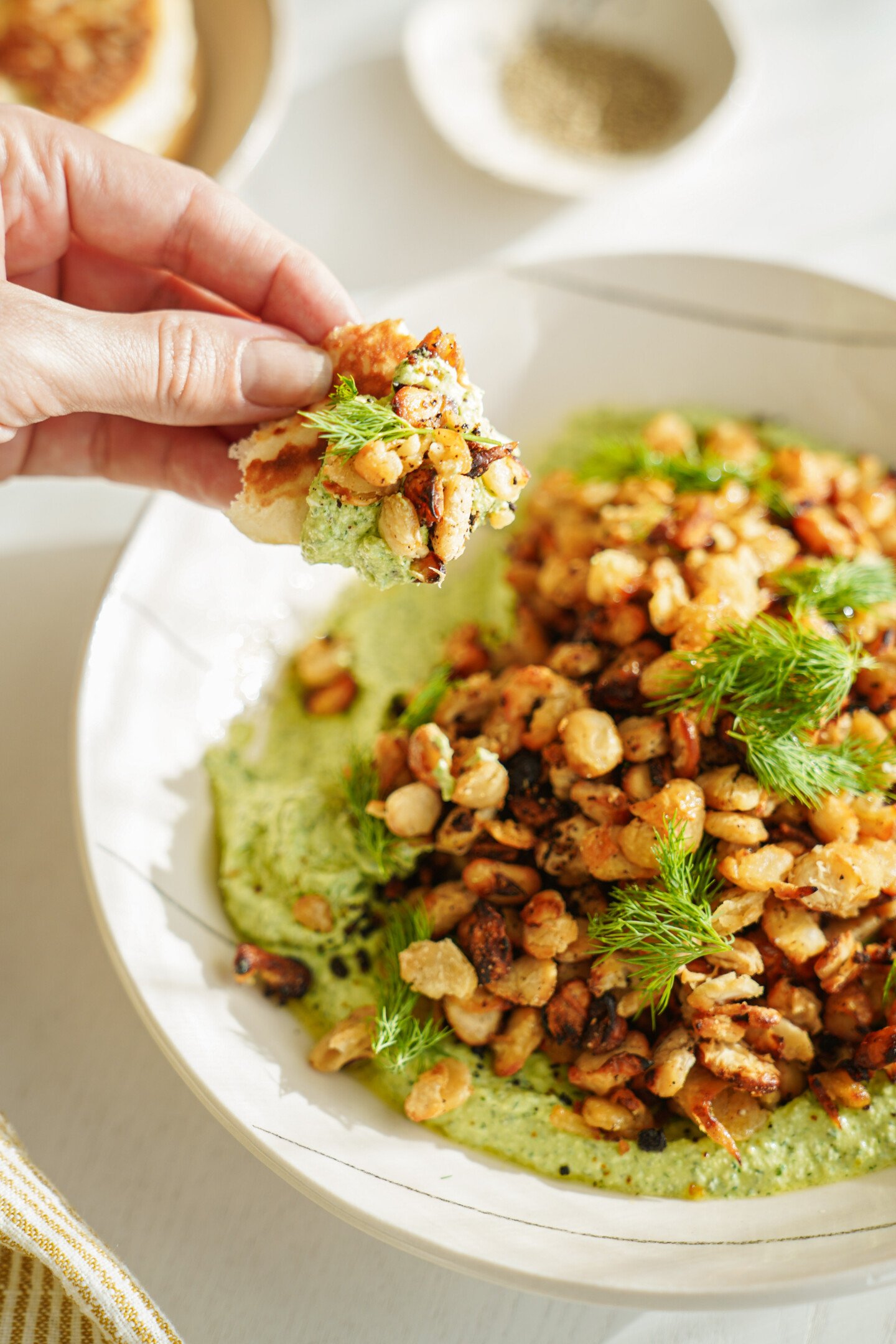A hand holds a piece of bread topped with creamy green dip, roasted chickpeas, and fresh herbs over a bowl filled with more dip, chickpeas, and herbs. A small bowl of pepper is in the background.