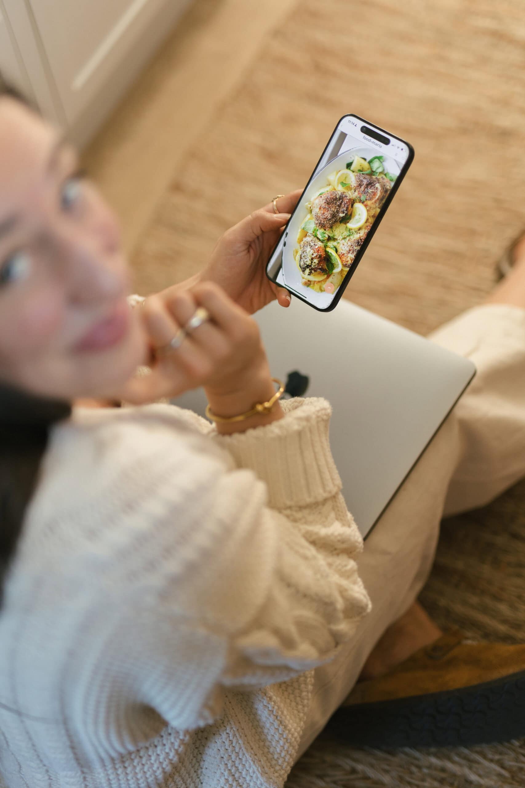 A person in a cream sweater sits on the floor holding a smartphone that displays a photo of a plated meal with greens and sauce. The person is smiling and holding a closed laptop on their lap.