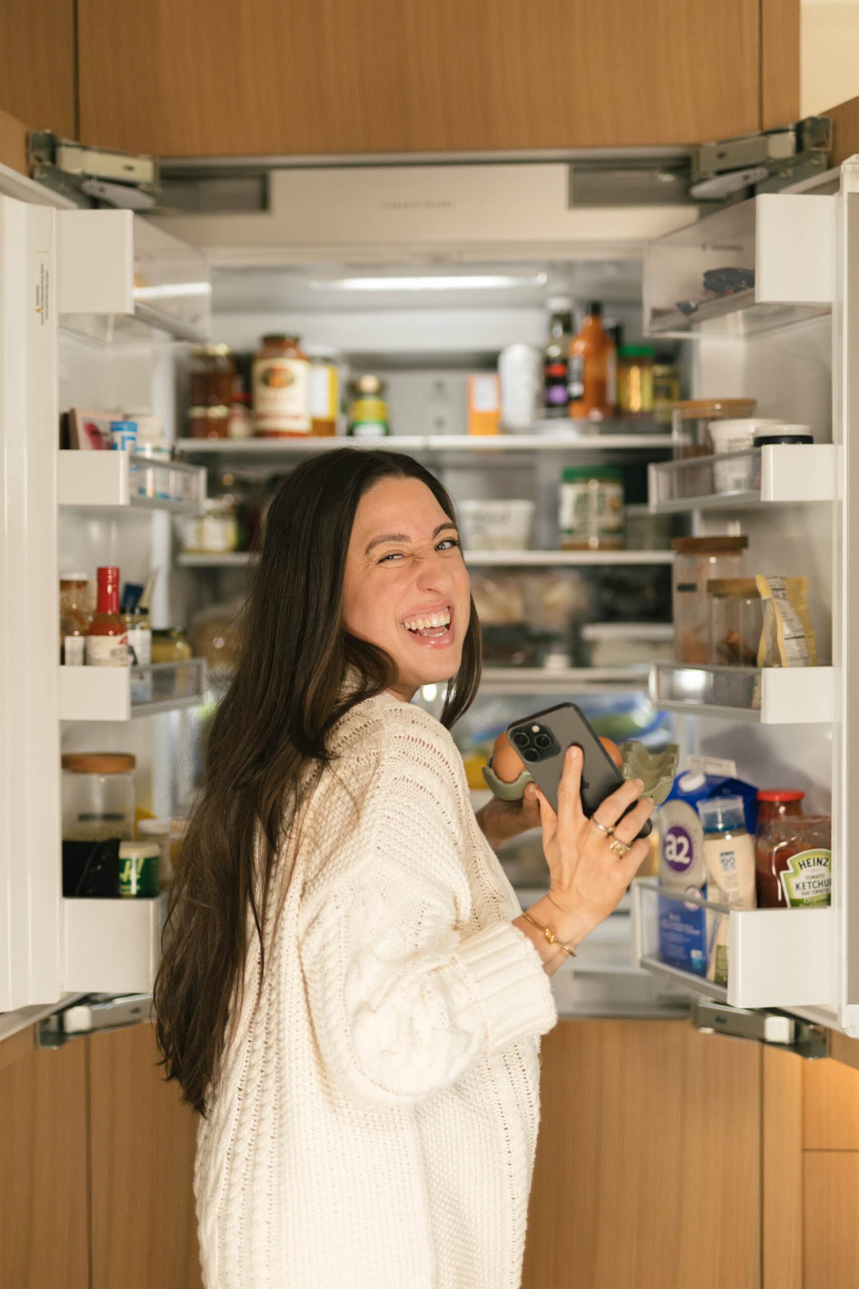 A woman with long dark hair, wearing a white sweater, stands smiling and winking in front of an open refrigerator, holding a smartphone in her hand. The fridge is stocked with various food items and drinks.
