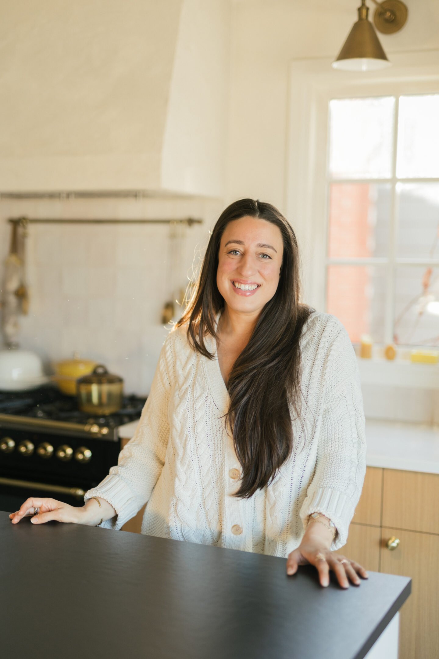 A woman with long brown hair and a cream-colored cable-knit sweater stands smiling in a bright kitchen with a stove and window in the background.