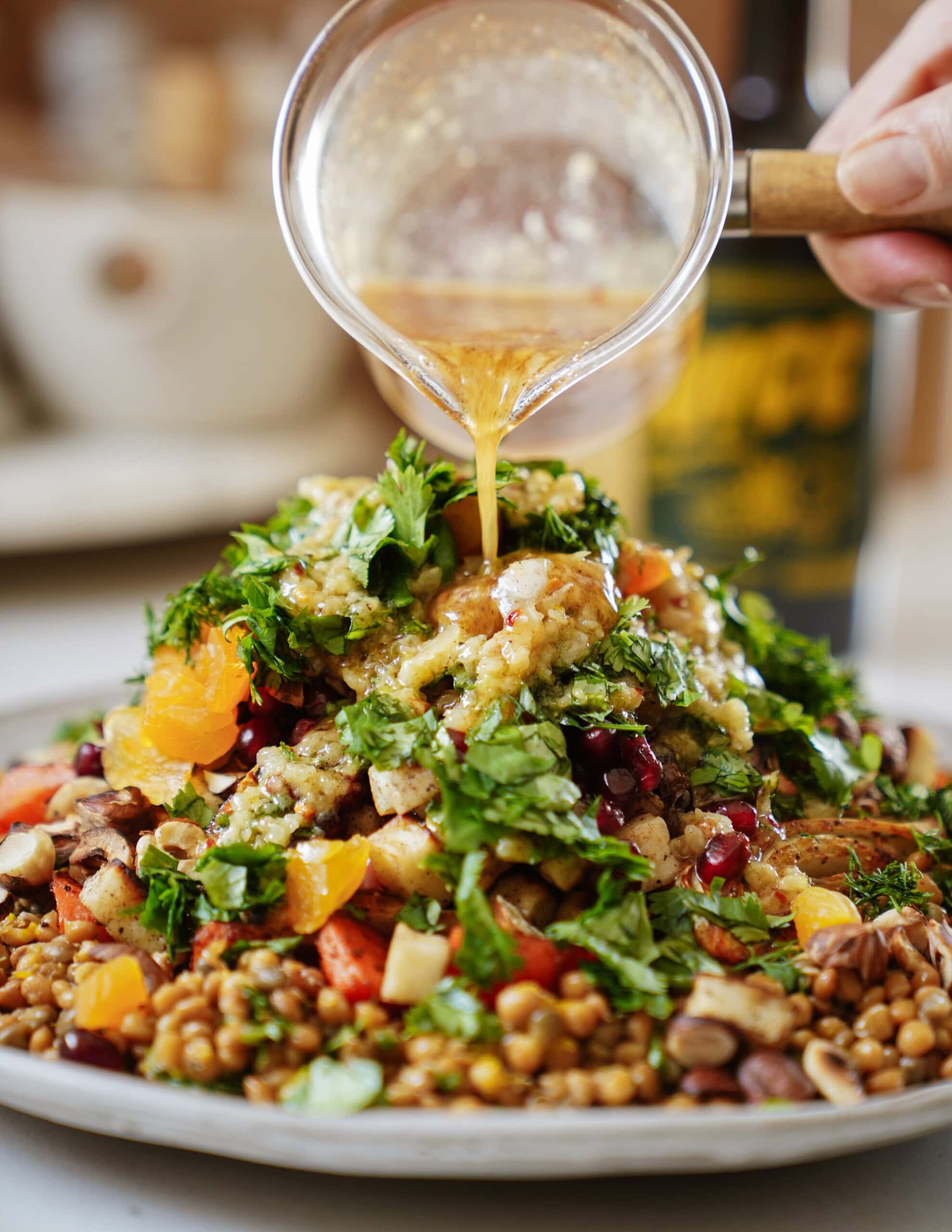 A hand pours dressing over a colorful lentil salad topped with chopped herbs, orange slices, pomegranate seeds, and nuts, all arranged on a plate.
