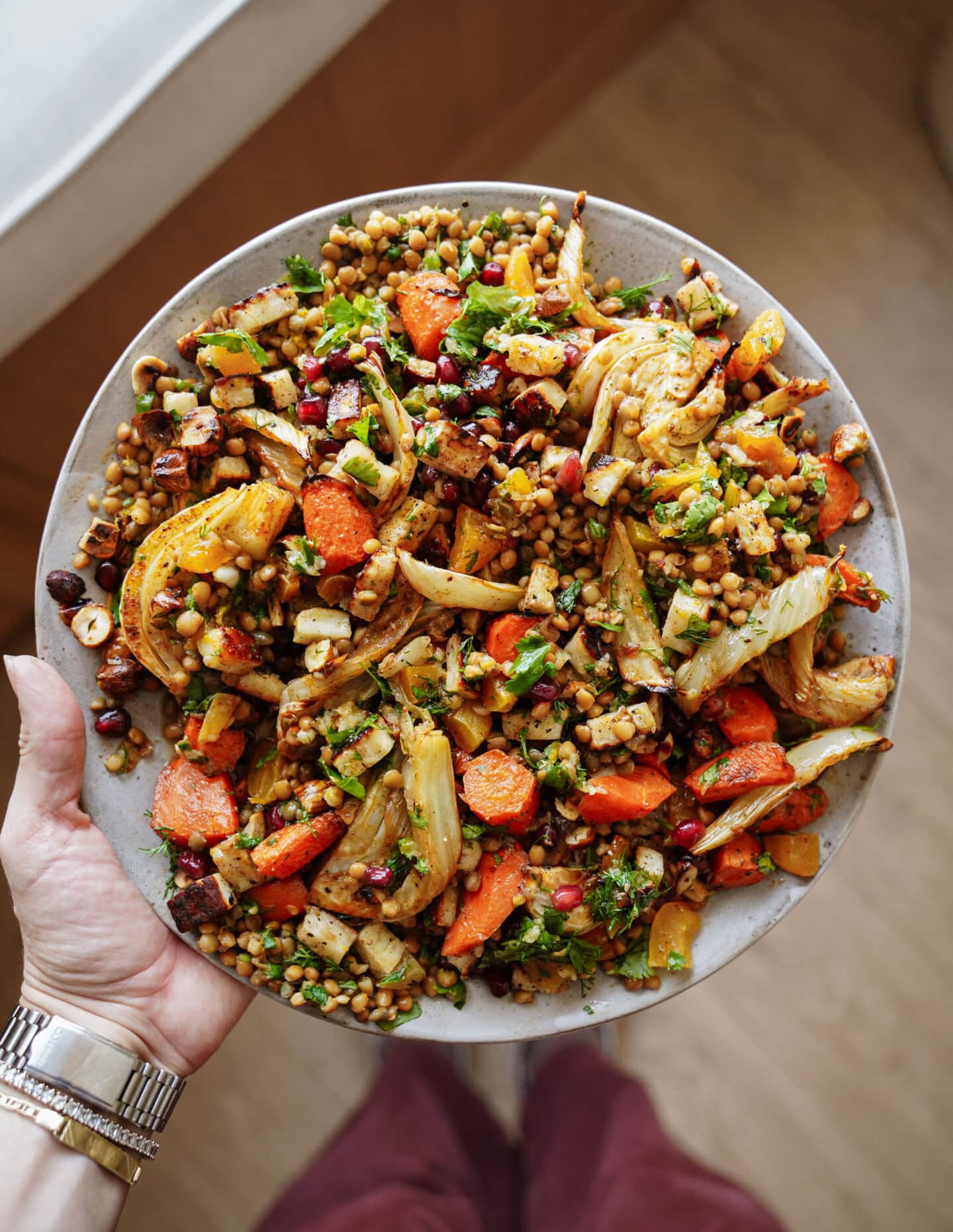 A hand holds a large plate filled with a colorful salad of roasted carrots, fennel, grains, chickpeas, fresh herbs, and pomegranate seeds on a wooden floor background.