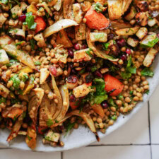 A close-up of a salad with roasted vegetables, pearl couscous, pomegranate seeds, leafy greens, and fresh herbs served on a white plate.