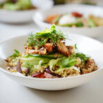A white bowl filled with quinoa salad topped with grilled chicken, sliced avocado, red onions, fresh herbs, and greens, with another bowl blurred in the background.