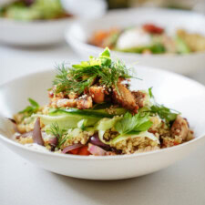A white bowl filled with quinoa salad topped with grilled chicken, sliced avocado, red onions, fresh herbs, and greens, with another bowl blurred in the background.