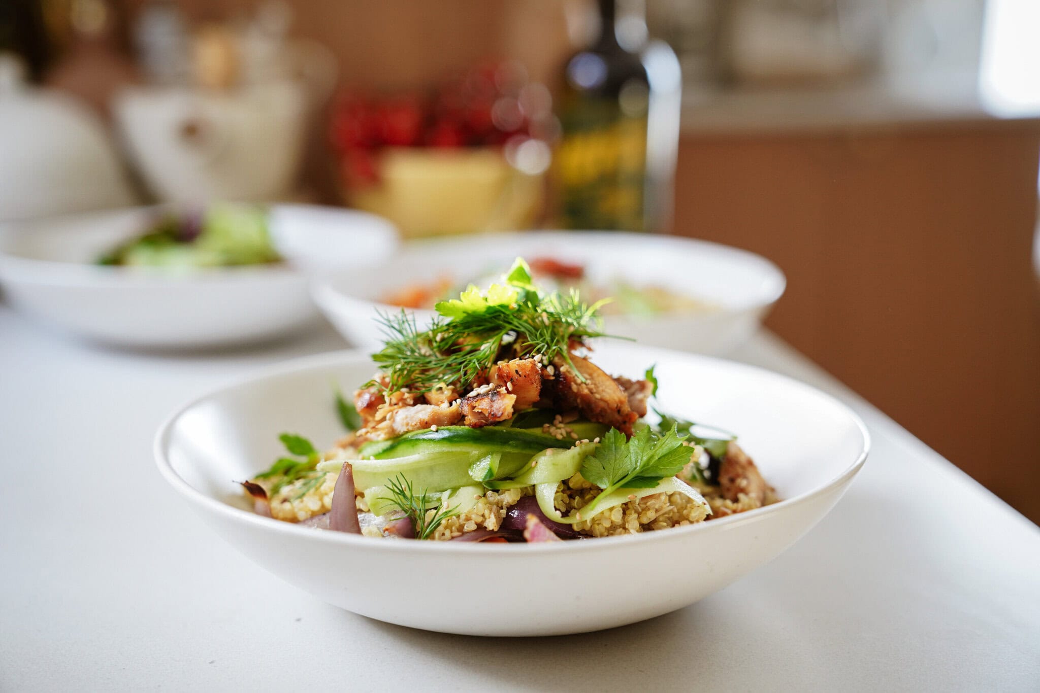 A white bowl filled with a colorful salad, topped with grilled chicken pieces, fresh parsley, dill, and snap peas, sits on a light-colored countertop. Other bowls and kitchen items are blurred in the background.