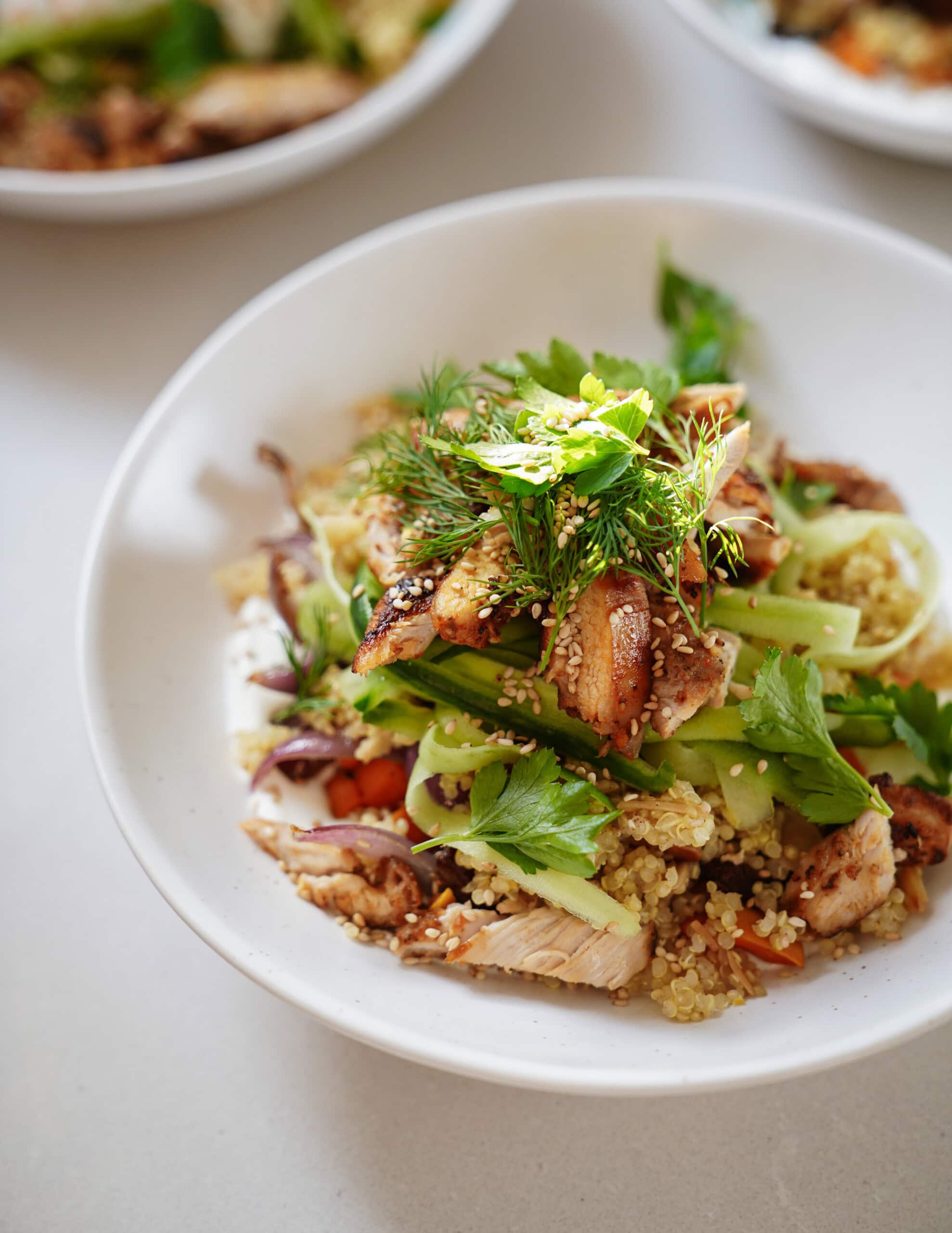 A bowl of quinoa salad topped with grilled chicken, fresh parsley, dill, celery slices, and sesame seeds, served in a white dish on a light-colored surface.