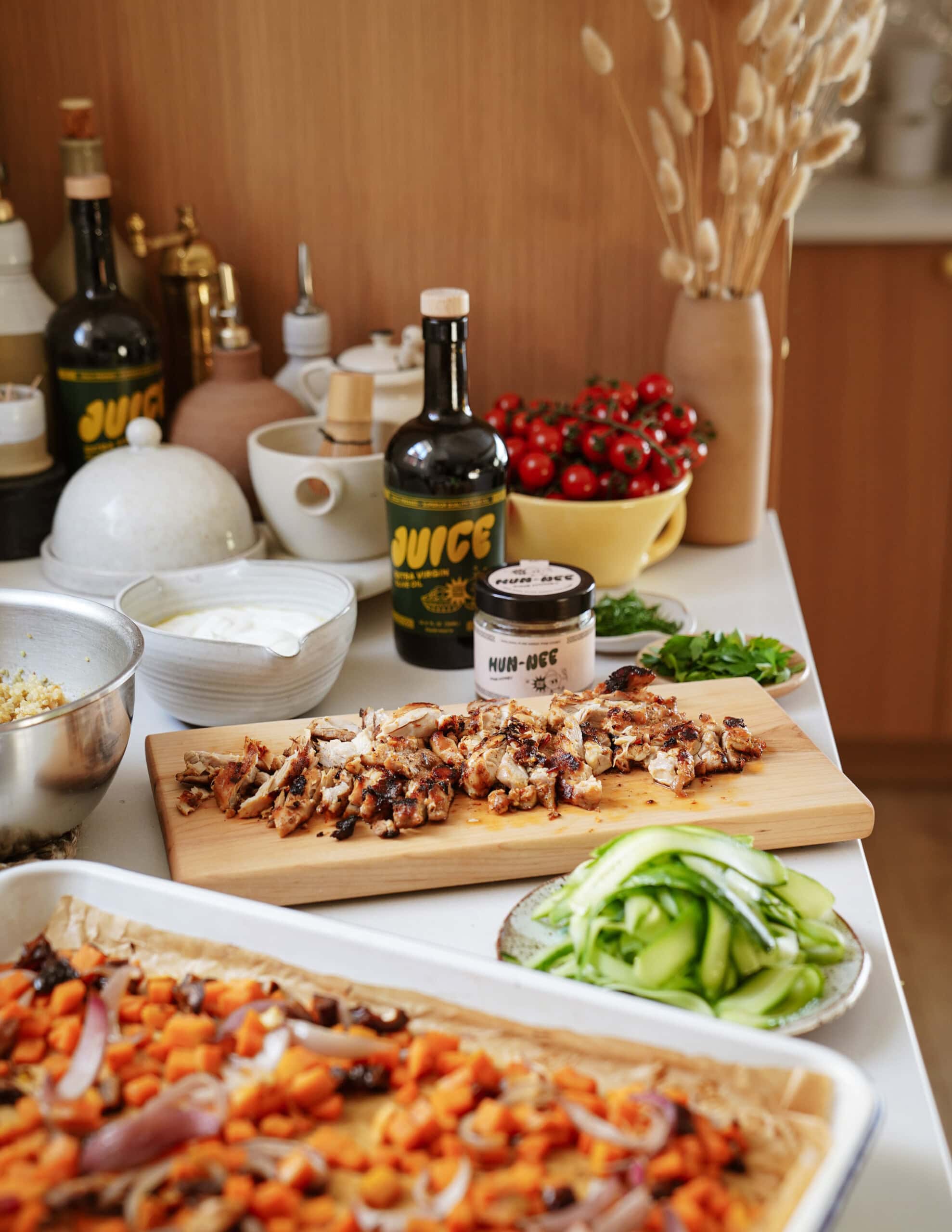 A kitchen counter displays grilled chicken on a cutting board, fresh sliced cucumbers, chopped herbs, a tray of roasted vegetables, cherry tomatoes, bottles of juice, and various bowls with ingredients.