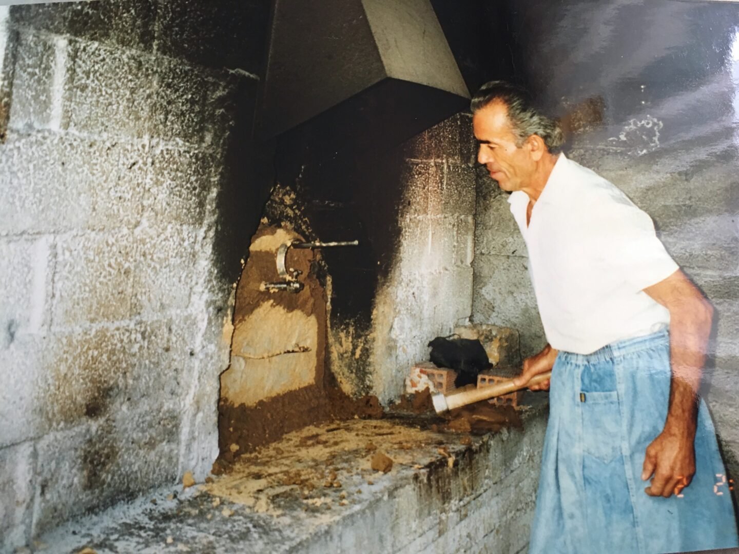 An older man in a white shirt and blue pants uses a wooden tool to tend to an outdoor oven built from concrete blocks, with soot and debris visible around the ovens opening.