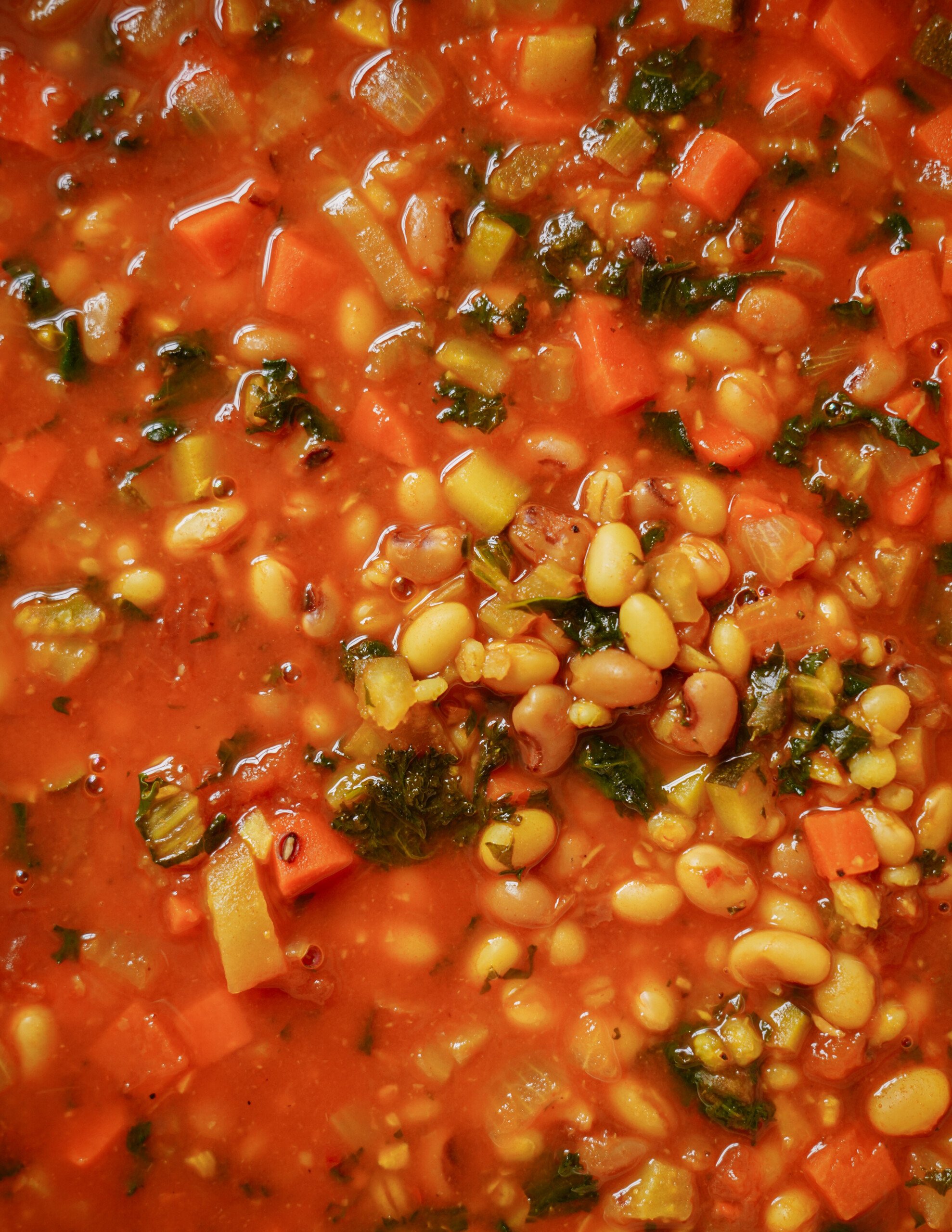 A close-up of a hearty vegetable soup featuring chopped carrots, leafy greens, white beans, and other vegetables in a rich, red tomato-based broth.