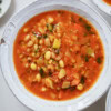 A white bowl filled with hearty bean soup, featuring white beans, diced tomatoes, celery, and fresh herbs in a rich, red broth.