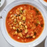A white bowl filled with hearty bean soup, featuring white beans, diced tomatoes, celery, and fresh herbs in a rich, red broth.