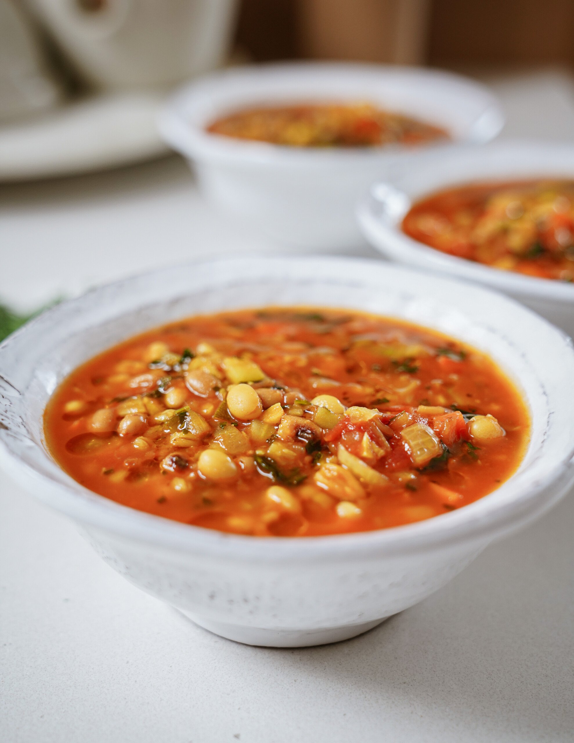 A close-up of a white bowl filled with vibrant vegetable soup containing beans, corn, tomatoes, and herbs, with two similar bowls blurred in the background.