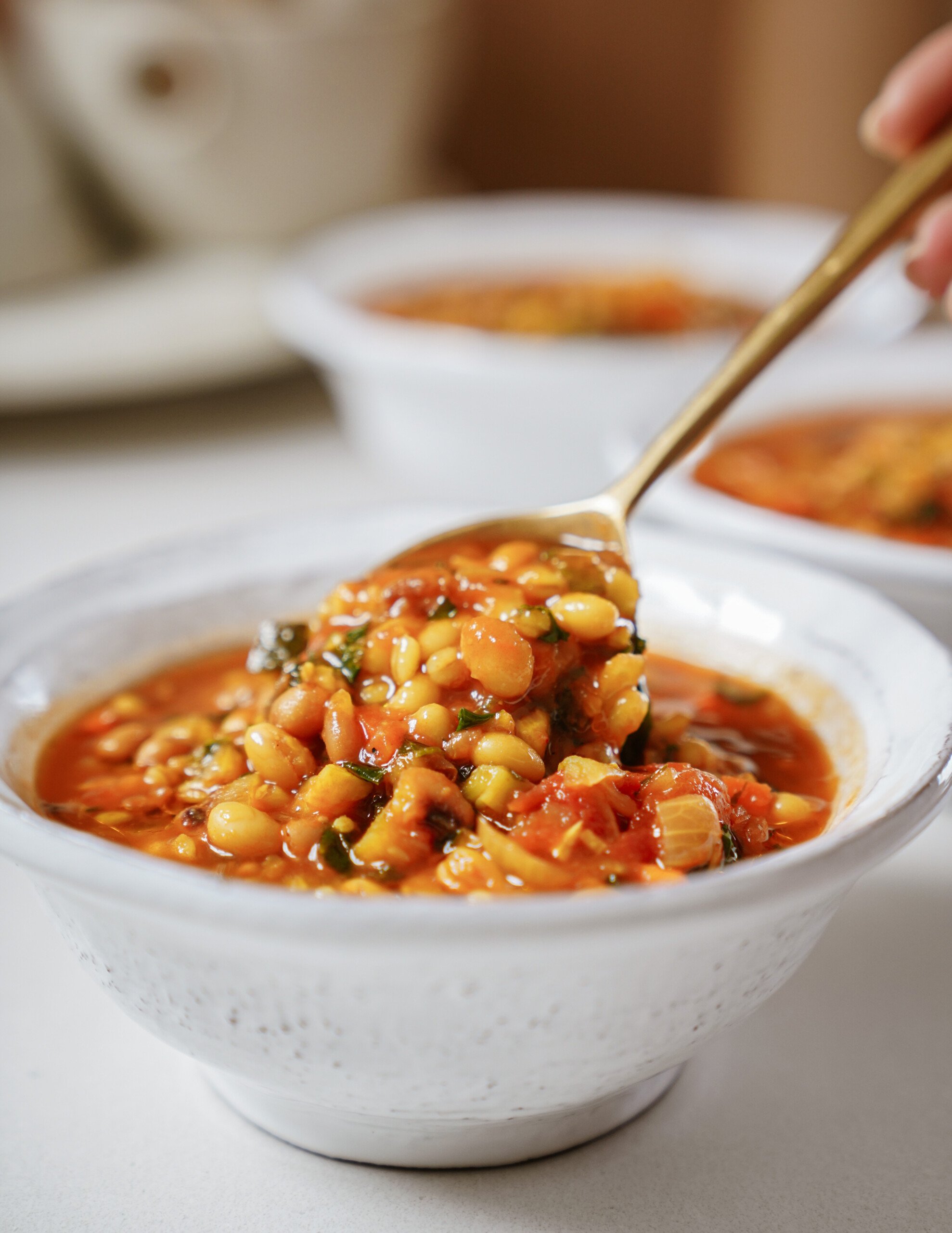 A close-up of a bowl of bean and vegetable soup with a spoon lifting a portion, showcasing beans, leafy greens, and a tomato-based broth. Two more bowls of soup are blurred in the background.