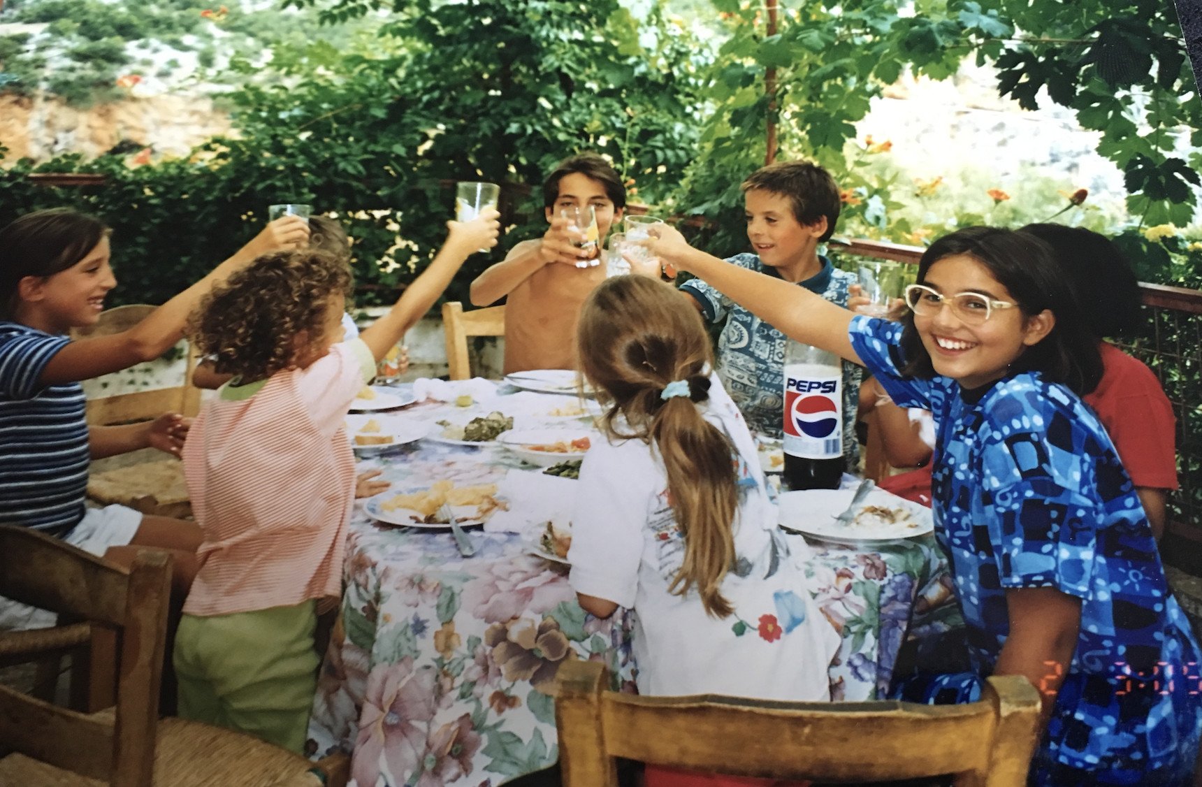 A group of six children sit around a table outdoors, smiling and raising glasses in a toast. The table is covered with a floral cloth, plates of food, and a large bottle of Pepsi. Lush greenery surrounds the scene.