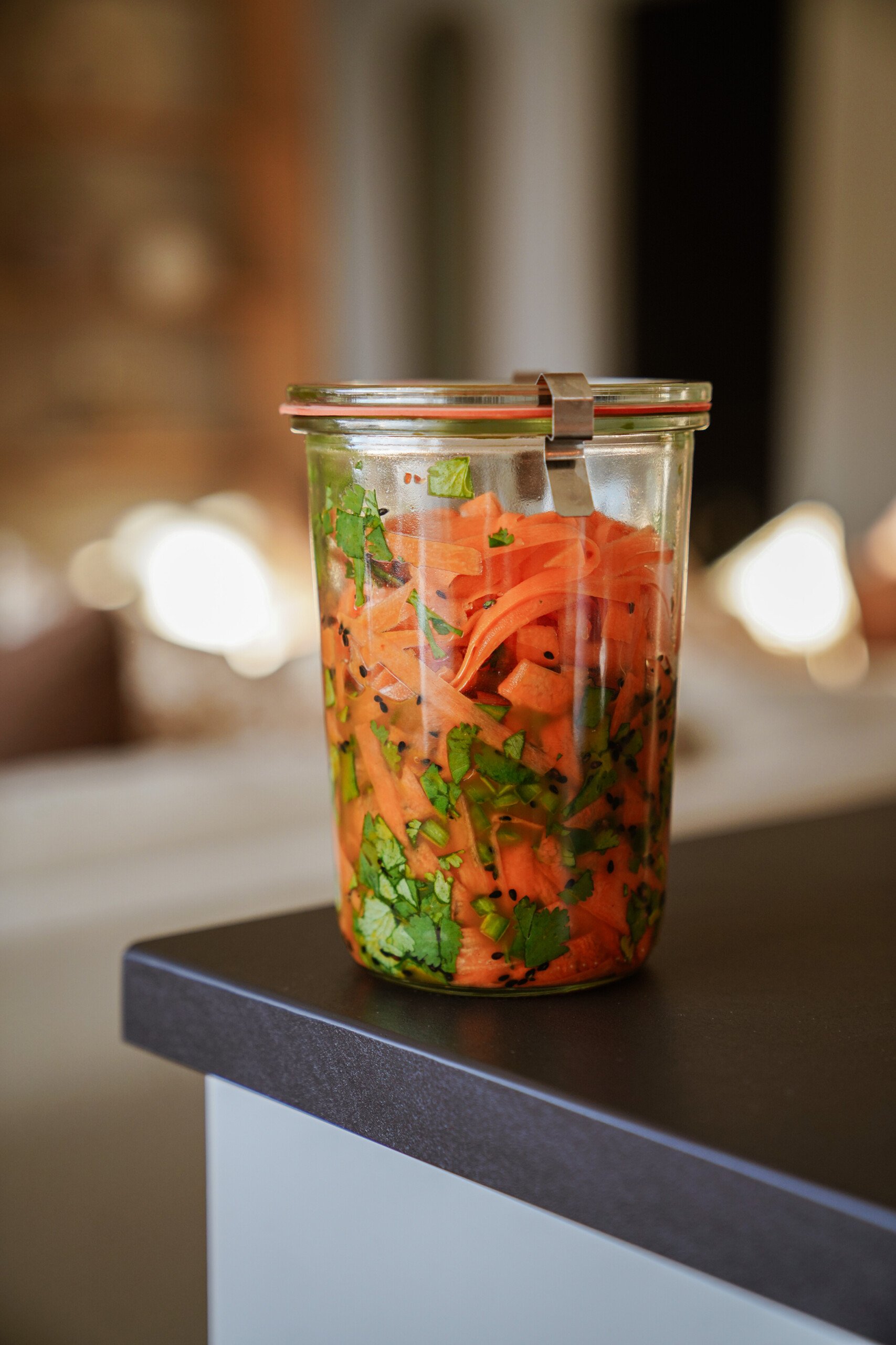 A glass jar filled with sliced carrots, chopped herbs, and spices sits on a dark countertop in a well-lit kitchen.