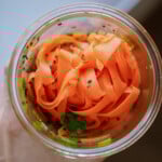 A glass jar filled with thinly sliced carrot ribbons, garnished with black sesame seeds and fresh green herbs, viewed from above.