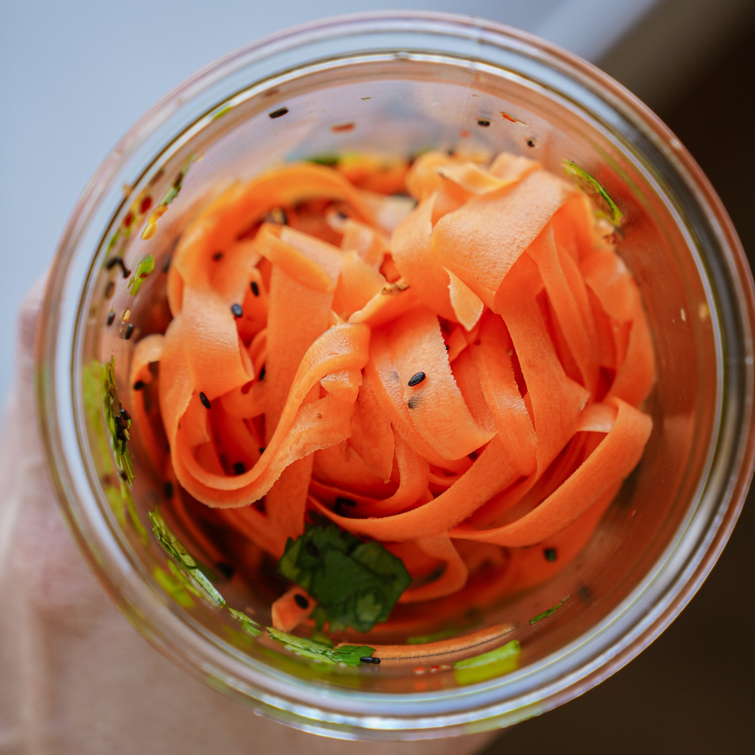 A glass jar filled with thinly sliced carrot ribbons, garnished with black sesame seeds and fresh green herbs, viewed from above.