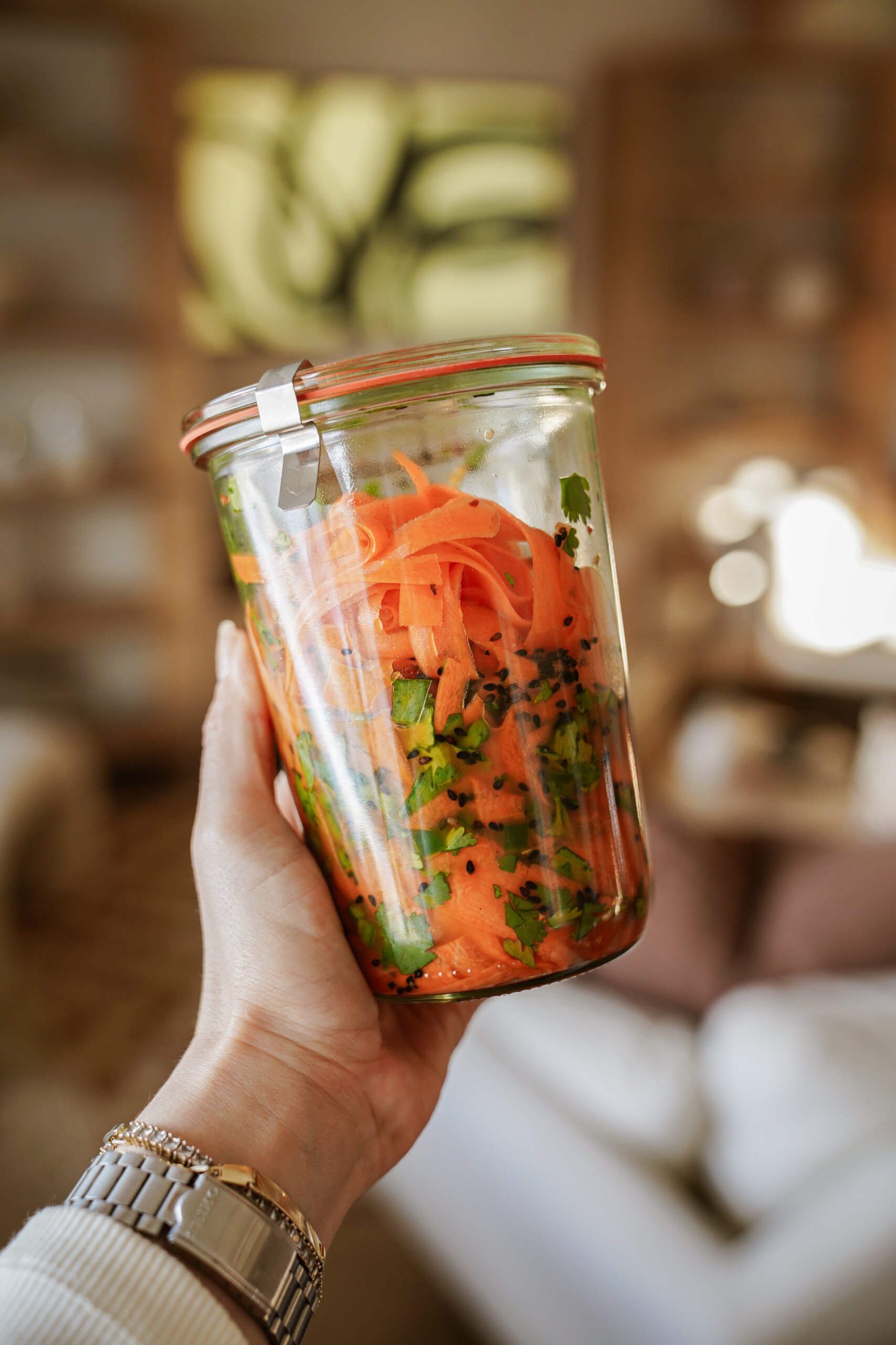 A hand holding a glass jar filled with spiralized carrots, chopped herbs, and seasonings. The background is a softly blurred indoor setting with warm lighting.