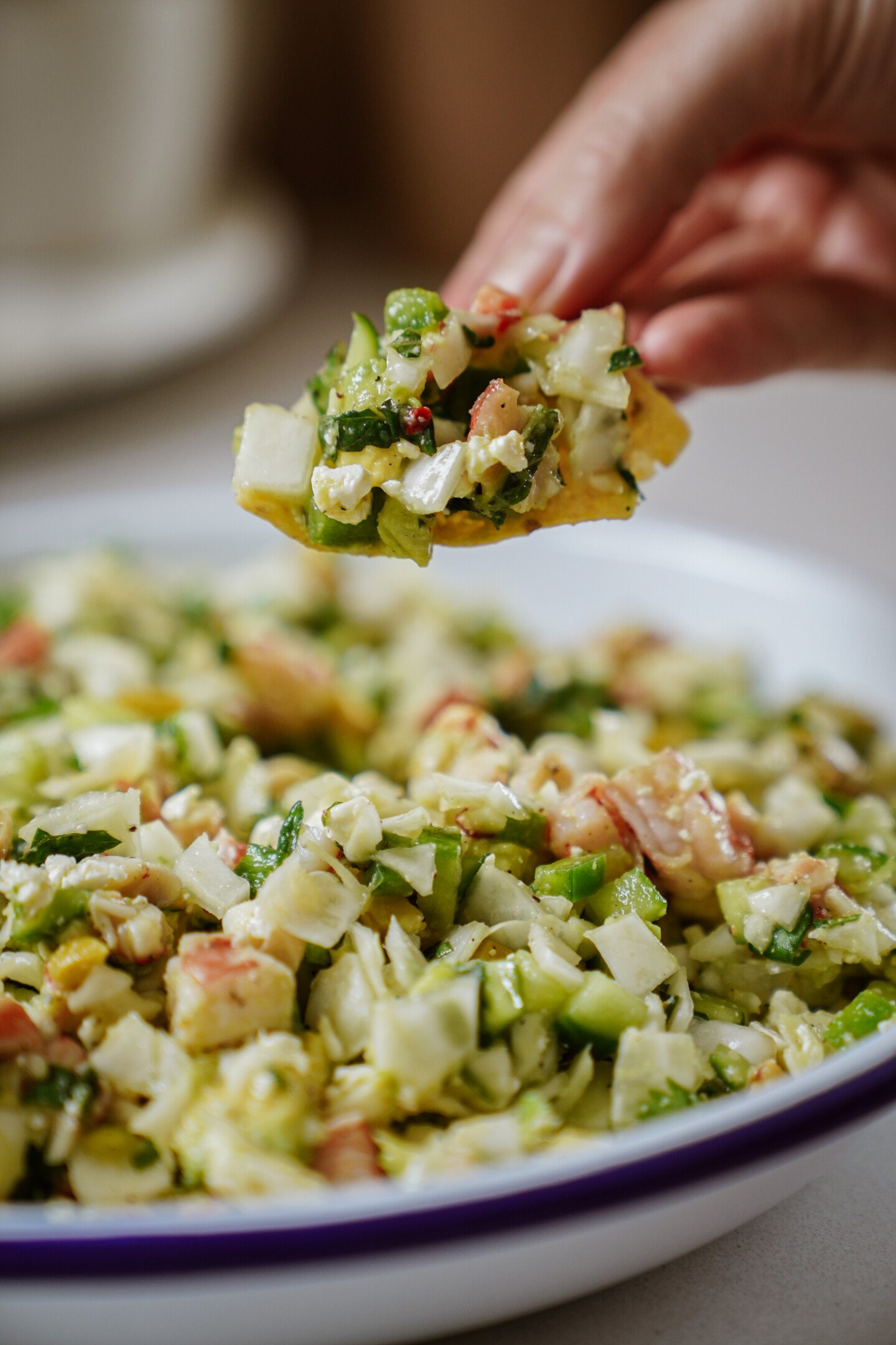 A hand holds a chip topped with a chunky vegetable and herb mixture, with a bowl of the same salad or salsa in the background.