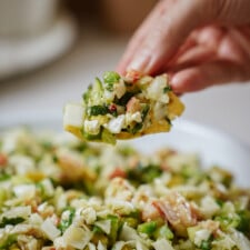 A hand holds a chip topped with a chunky mixture of chopped eggs, pickles, bacon, herbs, and vegetables over a plate filled with the same salad.