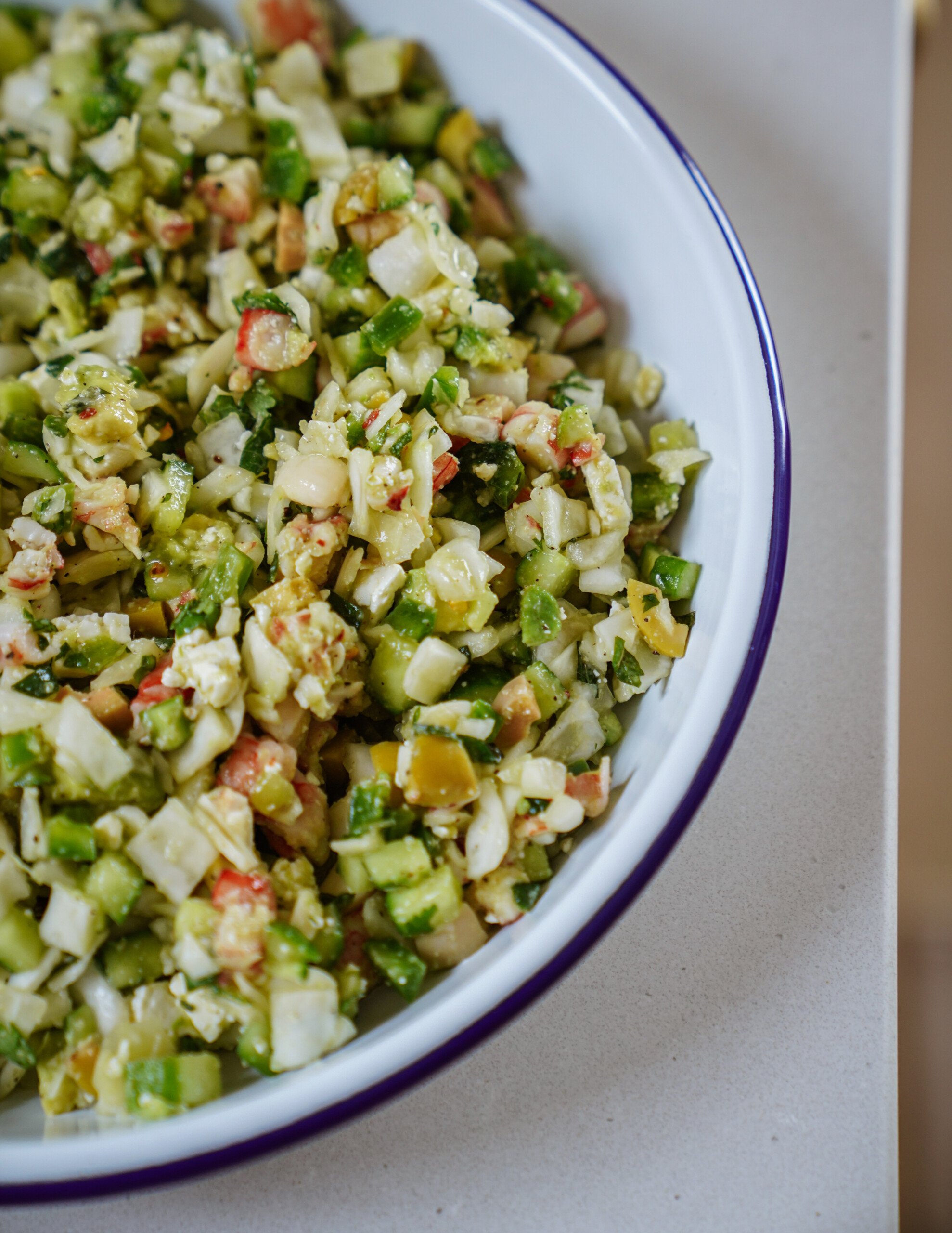 A close-up of a white bowl with a blue rim, filled with a finely chopped vegetable salad including cucumbers, peppers, onions, and other colorful ingredients, placed on a light countertop.