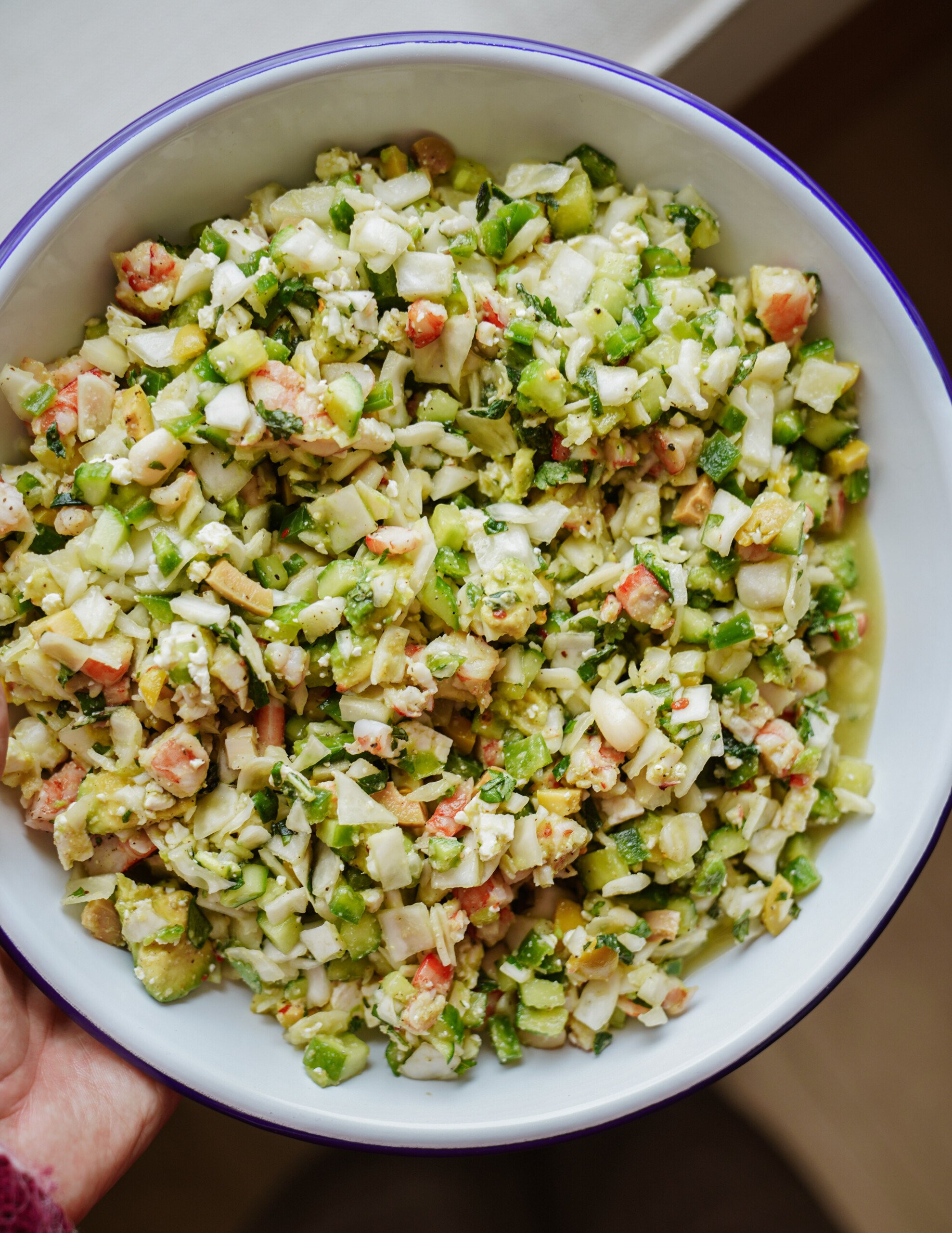 A bowl filled with a finely chopped salad containing green vegetables, white cubes, and small pieces of red, all mixed together with herbs in a light dressing.