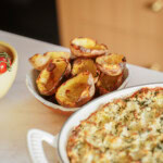 A bowl filled with crispy baked potato skins sits next to a white dish containing a cheesy, herb-topped casserole on a kitchen counter.