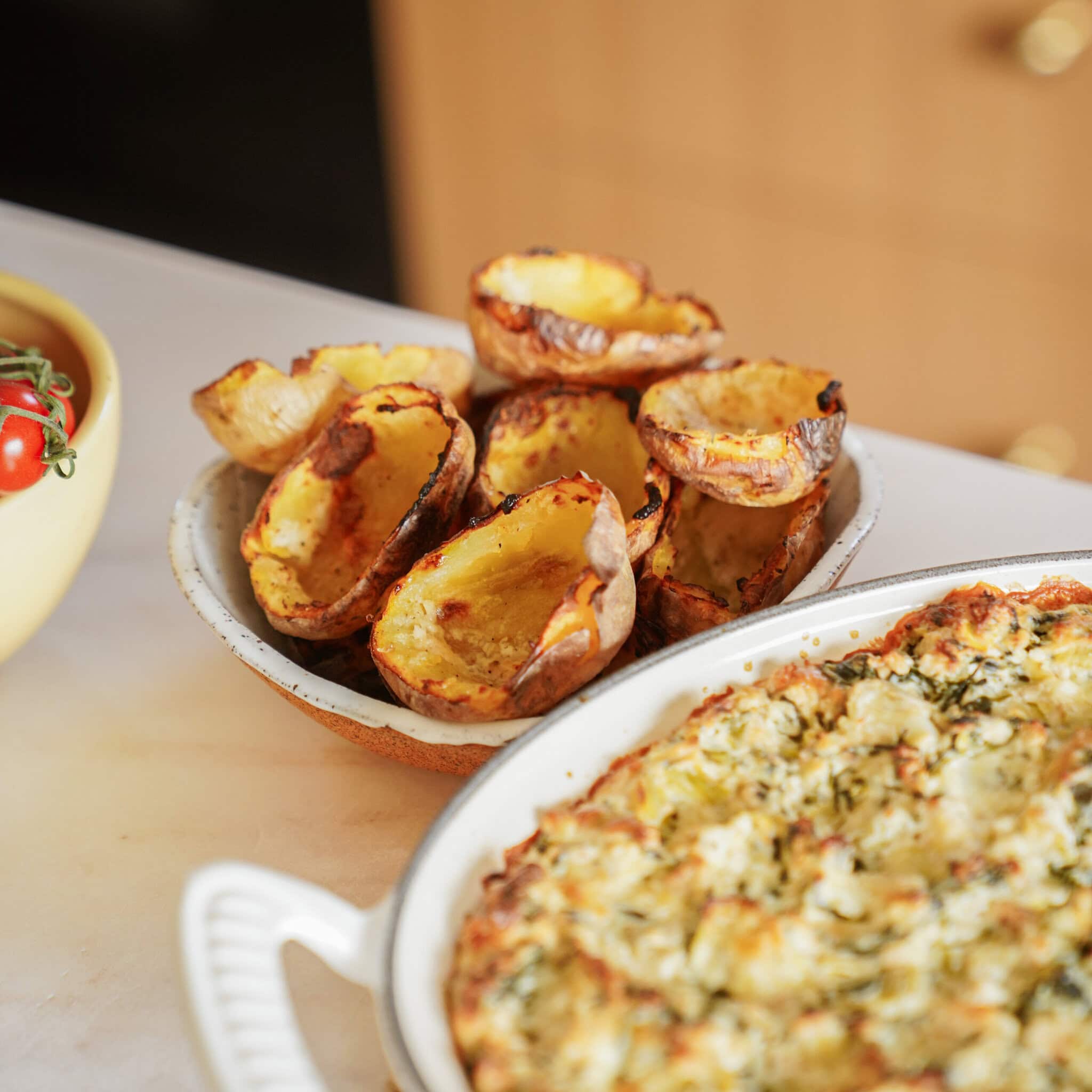 A bowl filled with crispy baked potato skins sits next to a white dish containing a cheesy, herb-topped casserole on a kitchen counter. - 1