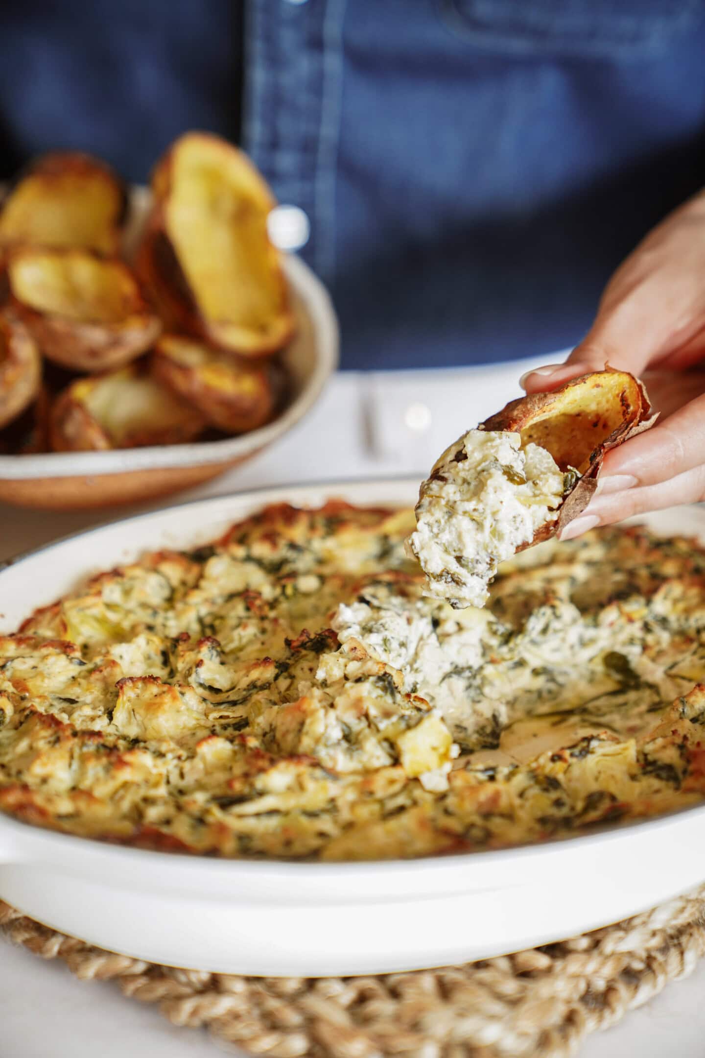 A hand scoops creamy spinach and artichoke dip from a white dish using a potato skin, with more potato skins in a bowl and a person in a blue shirt in the background.