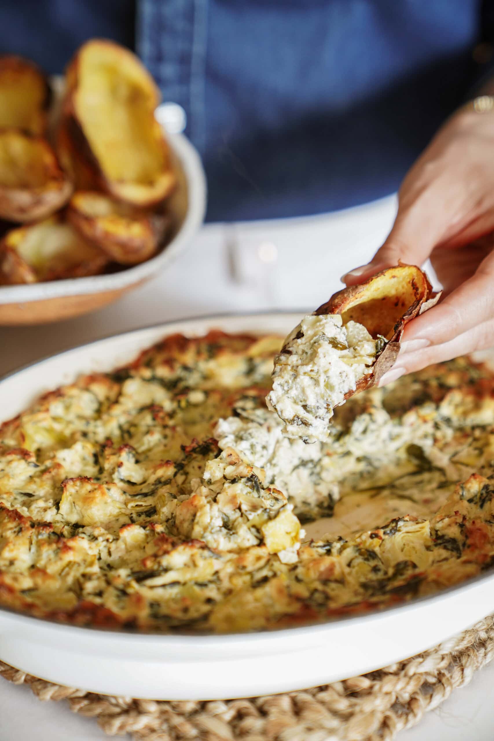 A hand scoops creamy spinach artichoke dip with a toasted bread slice from a white dish, with a bowl of toasted bread in the background.