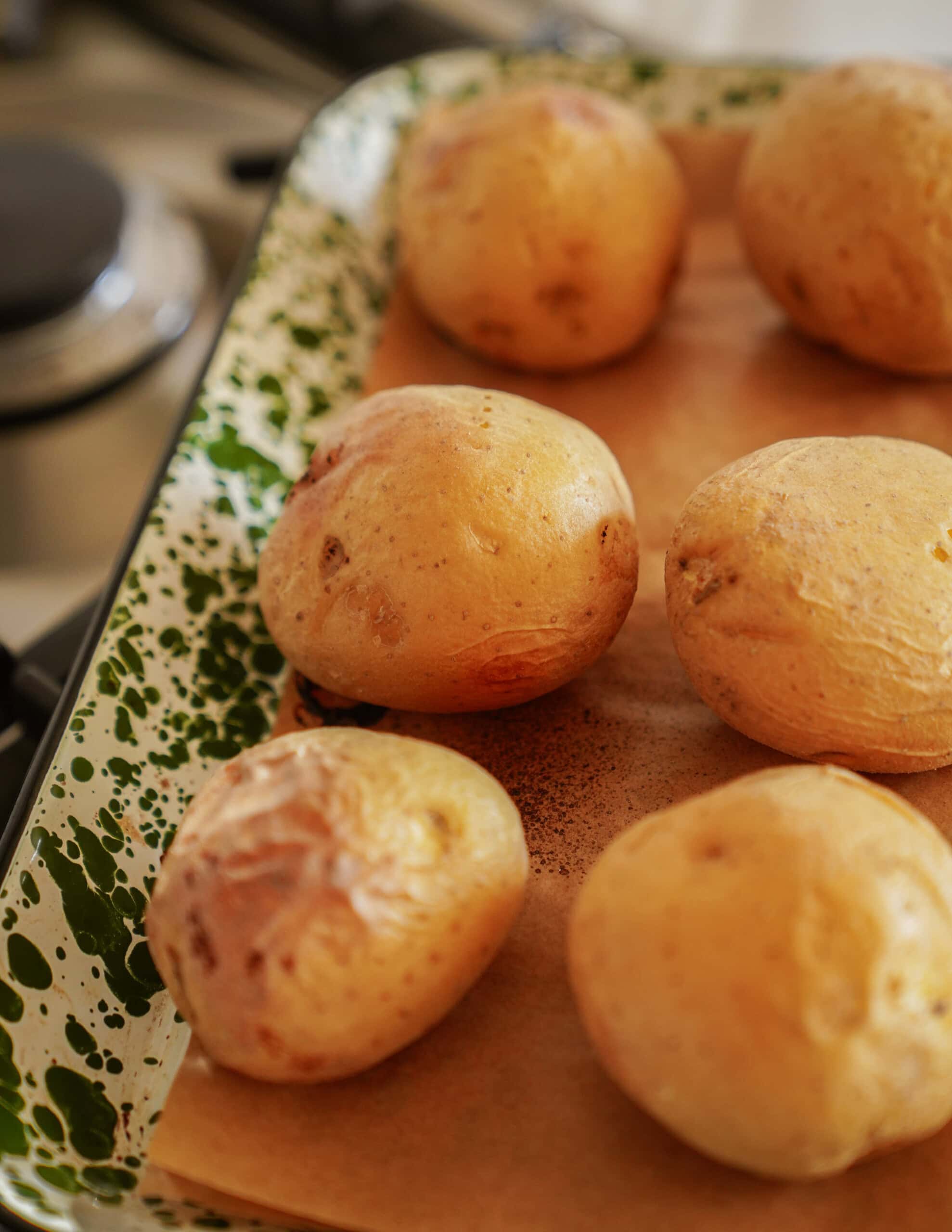 Six baked potatoes rest on a parchment-lined baking tray with a green and white speckled rim, on a kitchen countertop near a stovetop.