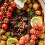 A dish with shredded roasted meat in the center, surrounded by roasted baby potatoes, vine tomatoes, fresh dill, lemon slices, and a dollop of white cheese or cream on top.