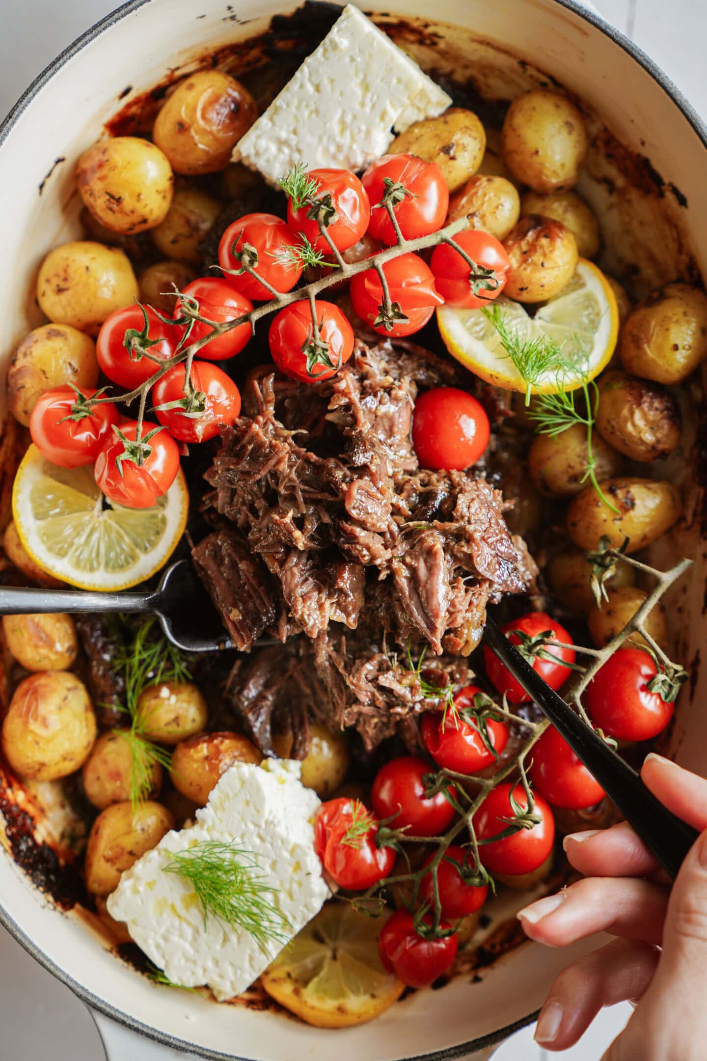 A hand holds a spoonful of shredded beef over a dish with roasted potatoes, whole cherry tomatoes on the vine, lemon slices, and blocks of feta cheese, all garnished with fresh dill.