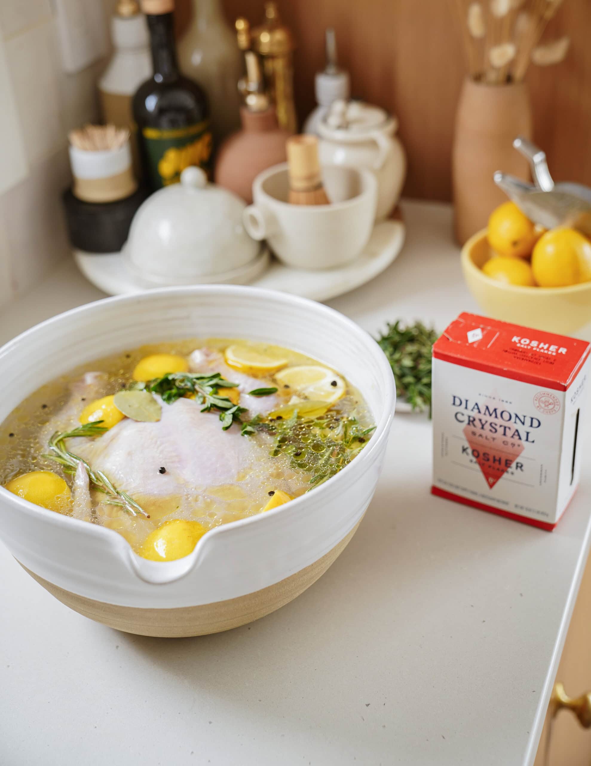 A large bowl with raw chicken, lemon slices, herbs, and spices in liquid on a kitchen counter. Nearby are a box of kosher salt, lemons, and various kitchen items.
