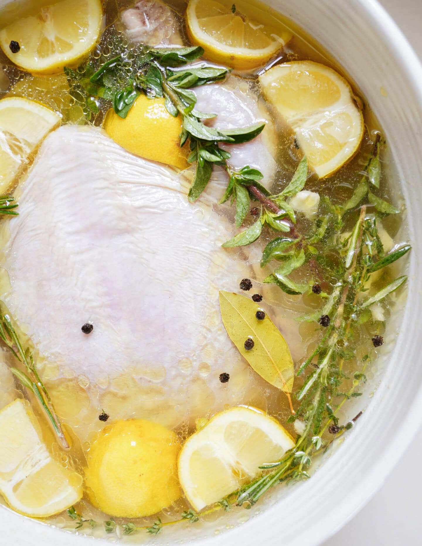 Raw chicken submerged in a marinade with lemon wedges, fresh herbs (thyme and oregano), bay leaves, black peppercorns, and liquid in a white bowl.
