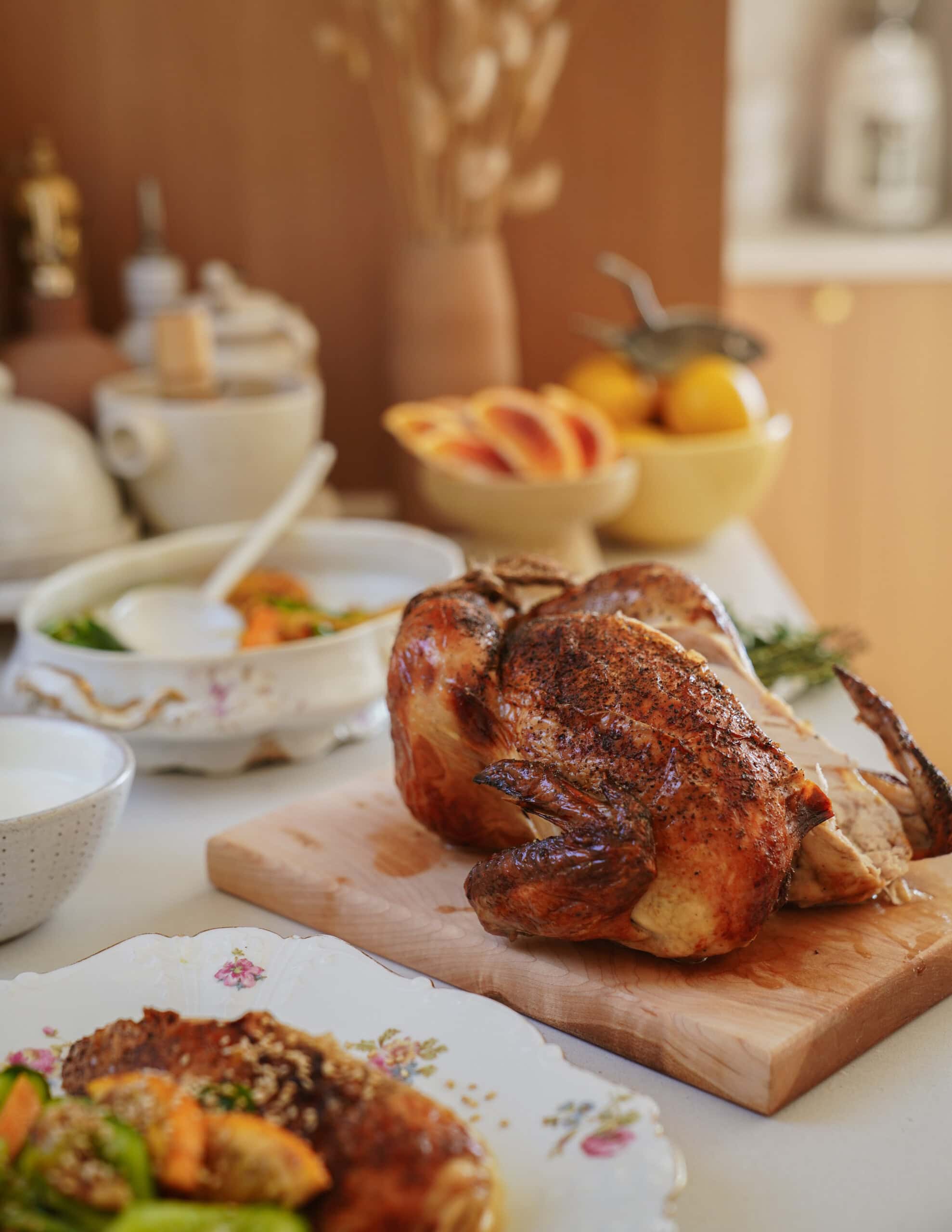 A roasted whole chicken sits on a wooden cutting board, surrounded by plates of vegetables and bowls on a table. In the background, there are fruit, dishes, and kitchen items with soft natural lighting.